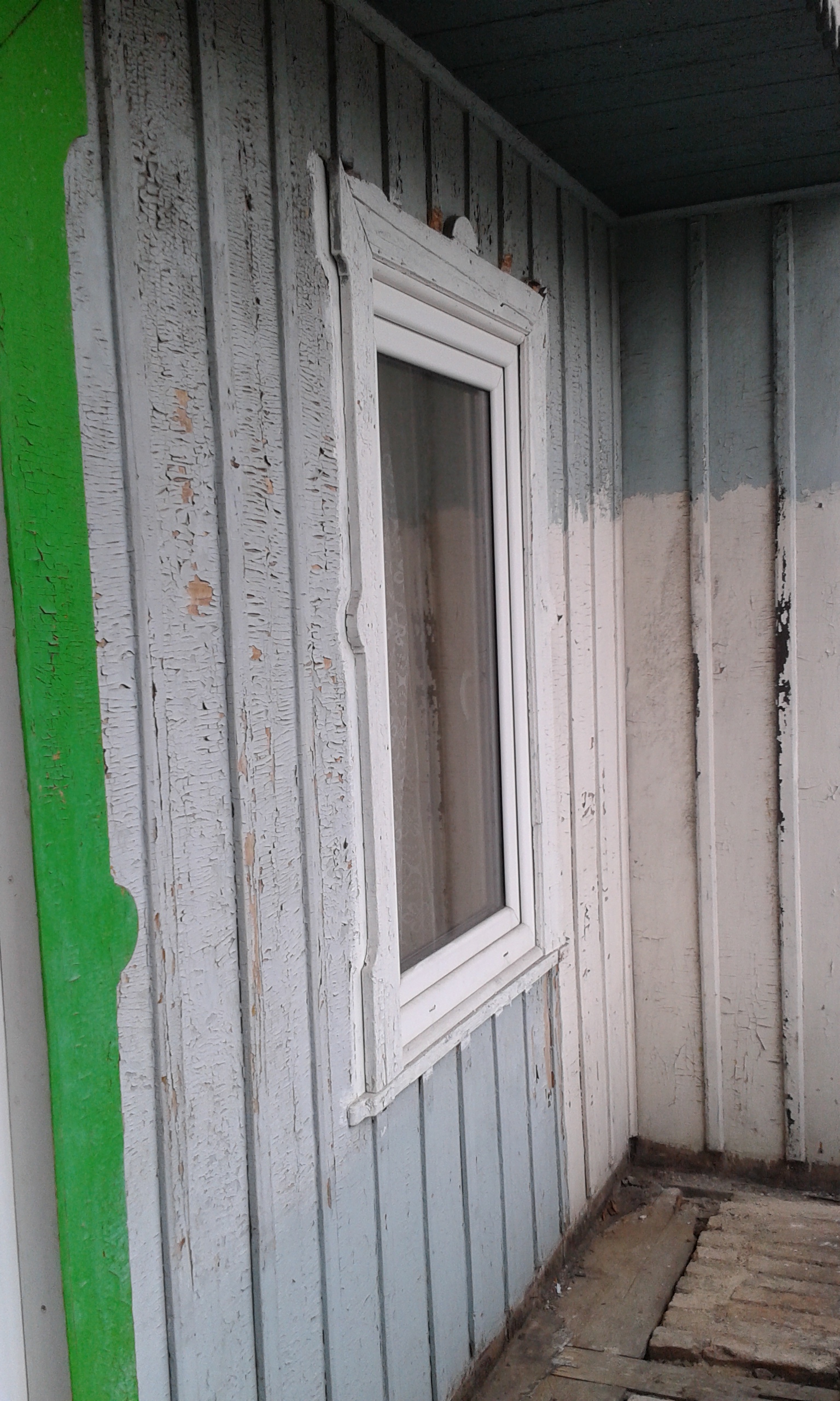 Stara drewniana ściana z pionowych desek w jasnoniebieskim kolorze, z białym oknem i fragmentem odsłoniętej podłogi, widok wnętrza przed remontem.