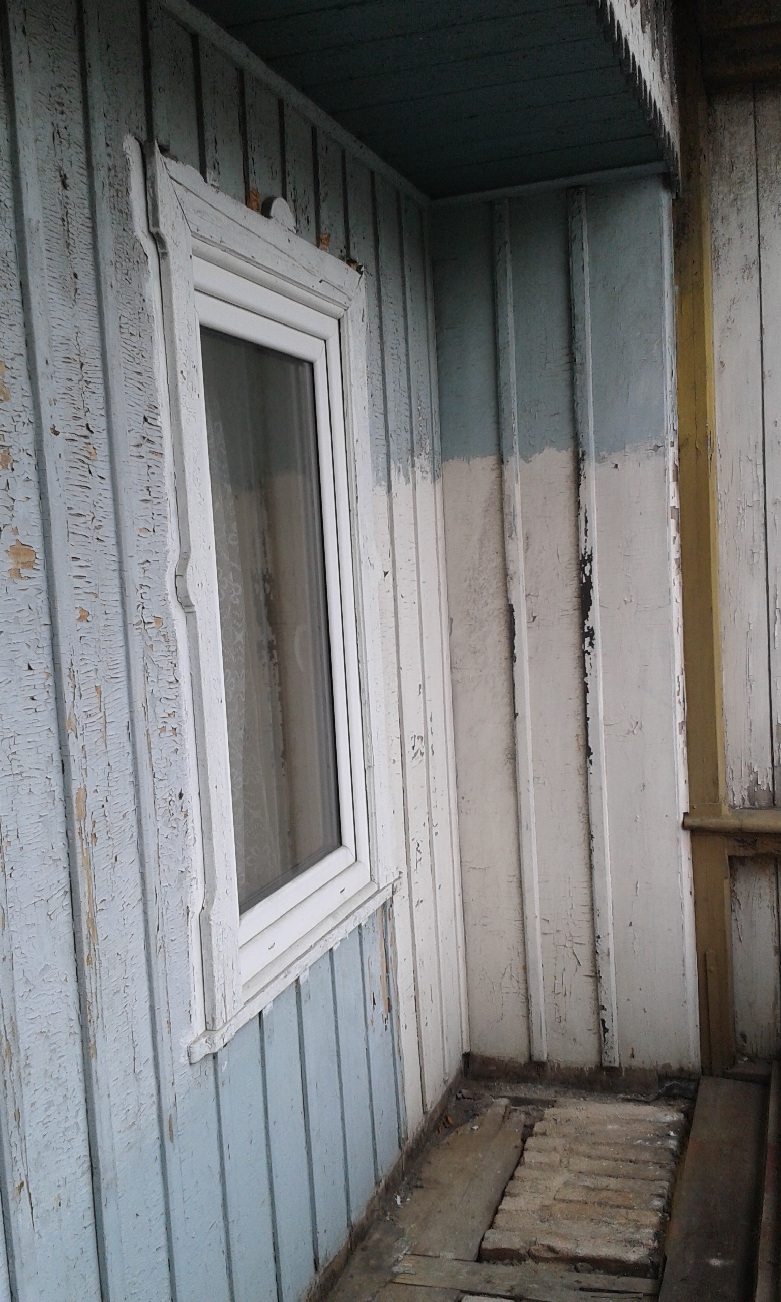 Stara drewniana ściana z pionowych desek w jasnoniebieskim kolorze, z widocznym oknem oraz fragmentem odsłoniętej podłogi przy ścianie podczas prac remontowych.