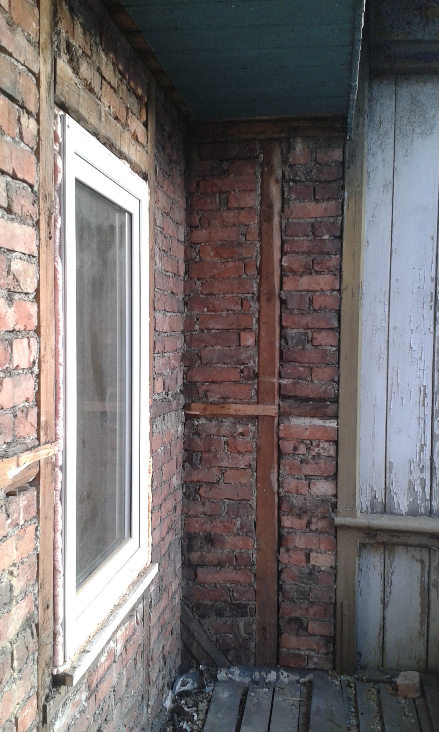 Ceglana wnęka balkonowa z białym oknem, widoczną drewnianą konstrukcją i surową ścianą podczas remontu.