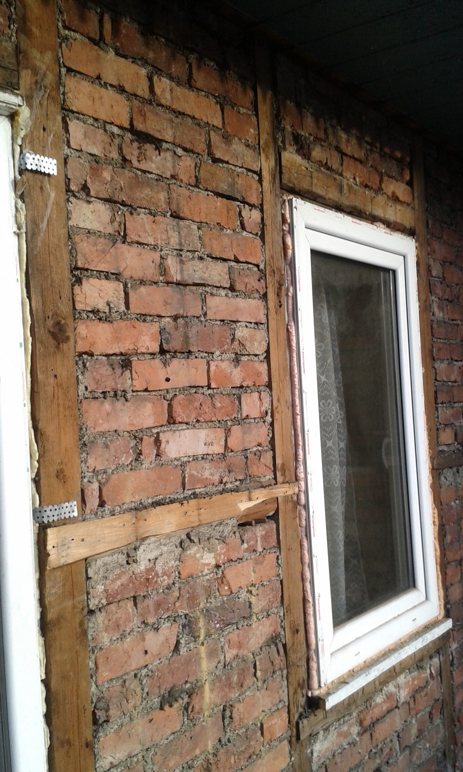 Ceglana ściana z zamontowanym oknem, odsłoniętymi belkami drewnianymi i widocznymi łączeniami konstrukcyjnymi podczas prac remontowych.