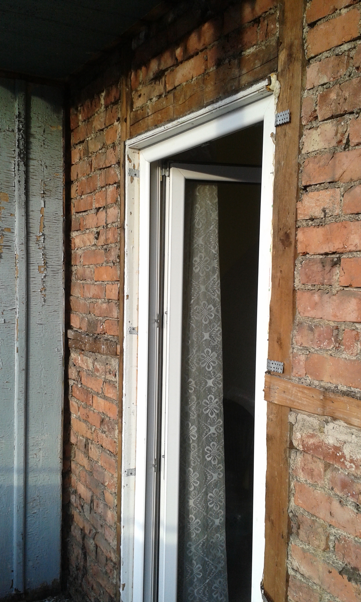 Otwór drzwiowy prowadzący na balkon z odsłoniętą ceglaną ścianą i widocznymi elementami drewnianej konstrukcji podczas prac remontowych.