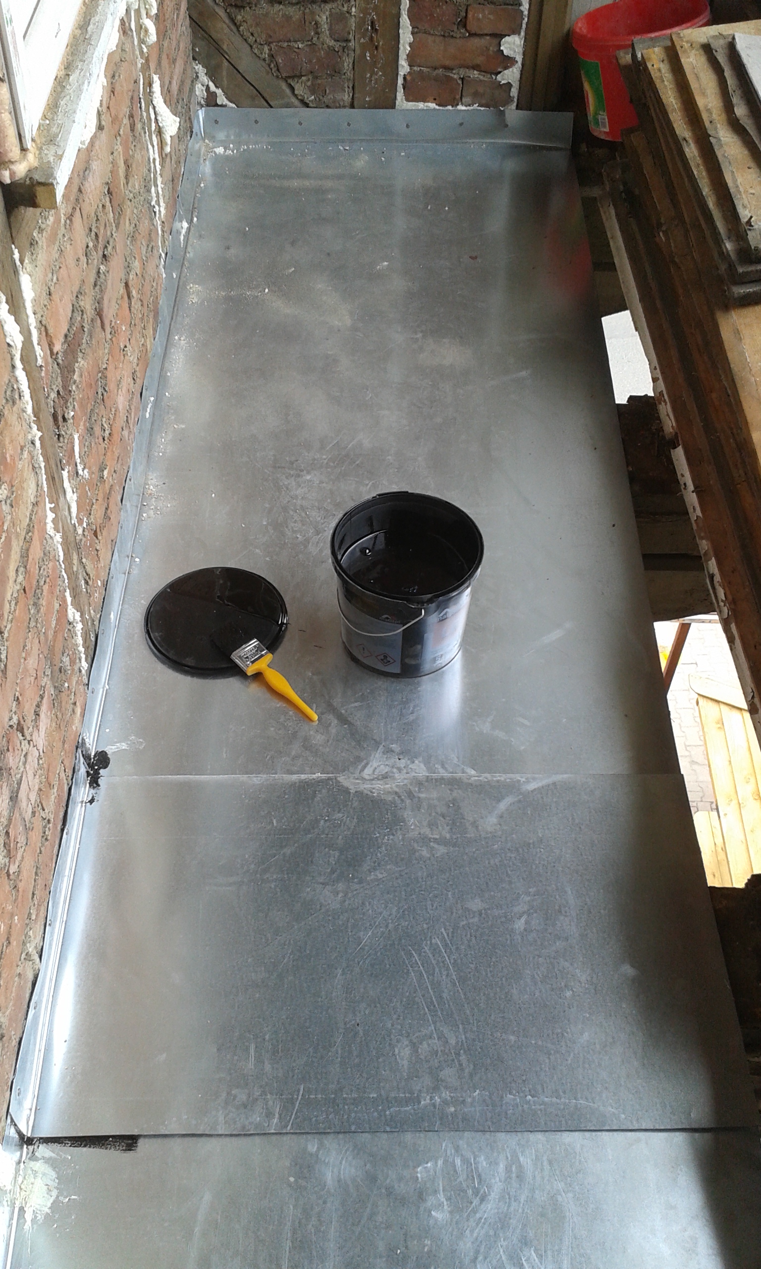 Arkusz blachy ułożony na podłodze balkonu jako zabezpieczenie w trakcie prac remontowych, obok puszka z masą i pędzel.