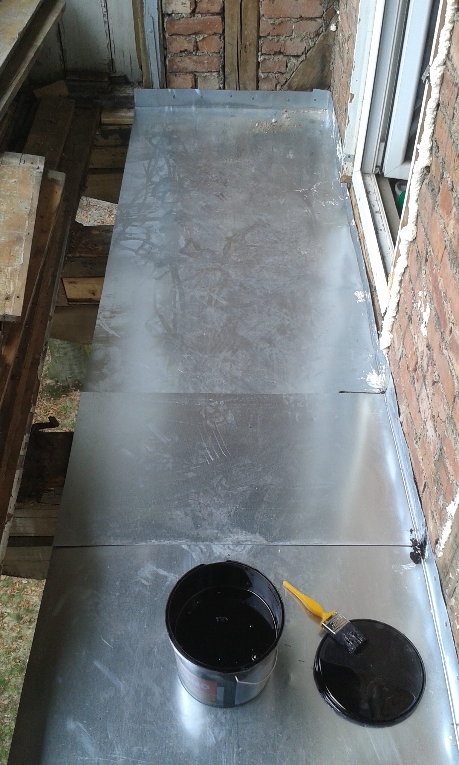 Podłoga balkonu zabezpieczona arkuszem blachy w trakcie prac remontowych, na blacie otwarta puszka z masą i pędzel.