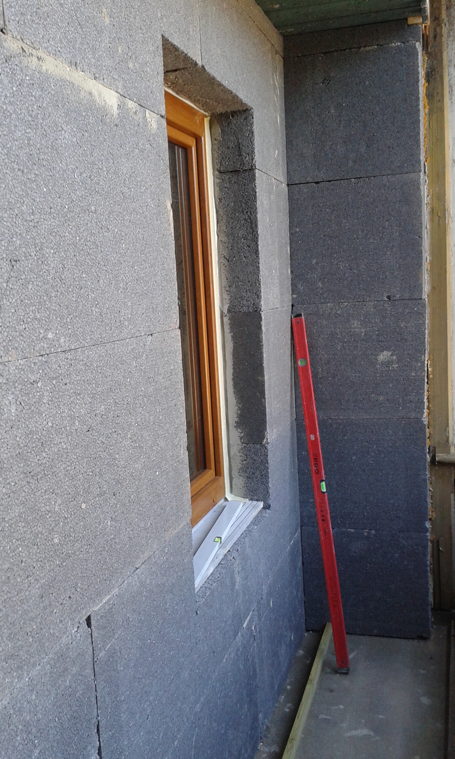 Wnęka okienna na balkonie w trakcie ocieplania, widoczne płyty grafitowego styropianu wokół okna oraz poziomica oparta o ścianę.