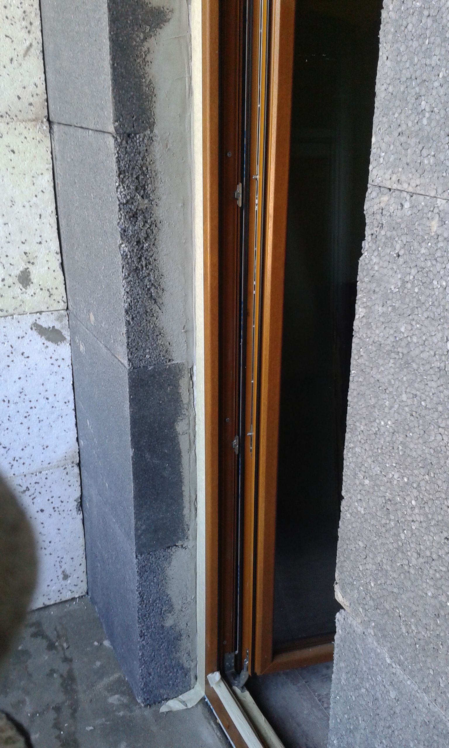 Detal ościeża balkonu z osadzonym oknem, widoczne warstwy grafitowego styropianu oraz kleju montażowego wokół ramy okiennej.