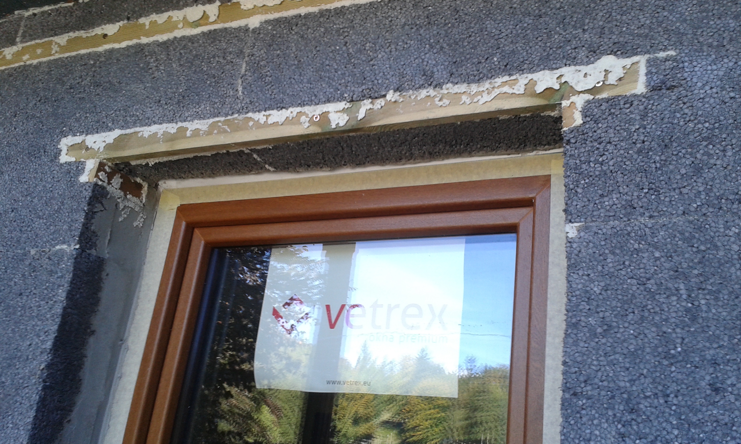 Okno balkonowe z brązową ramą osadzone w ścianie ocieplanej styropianem, z widocznymi szczelinami i resztkami kleju wokół nadproża.