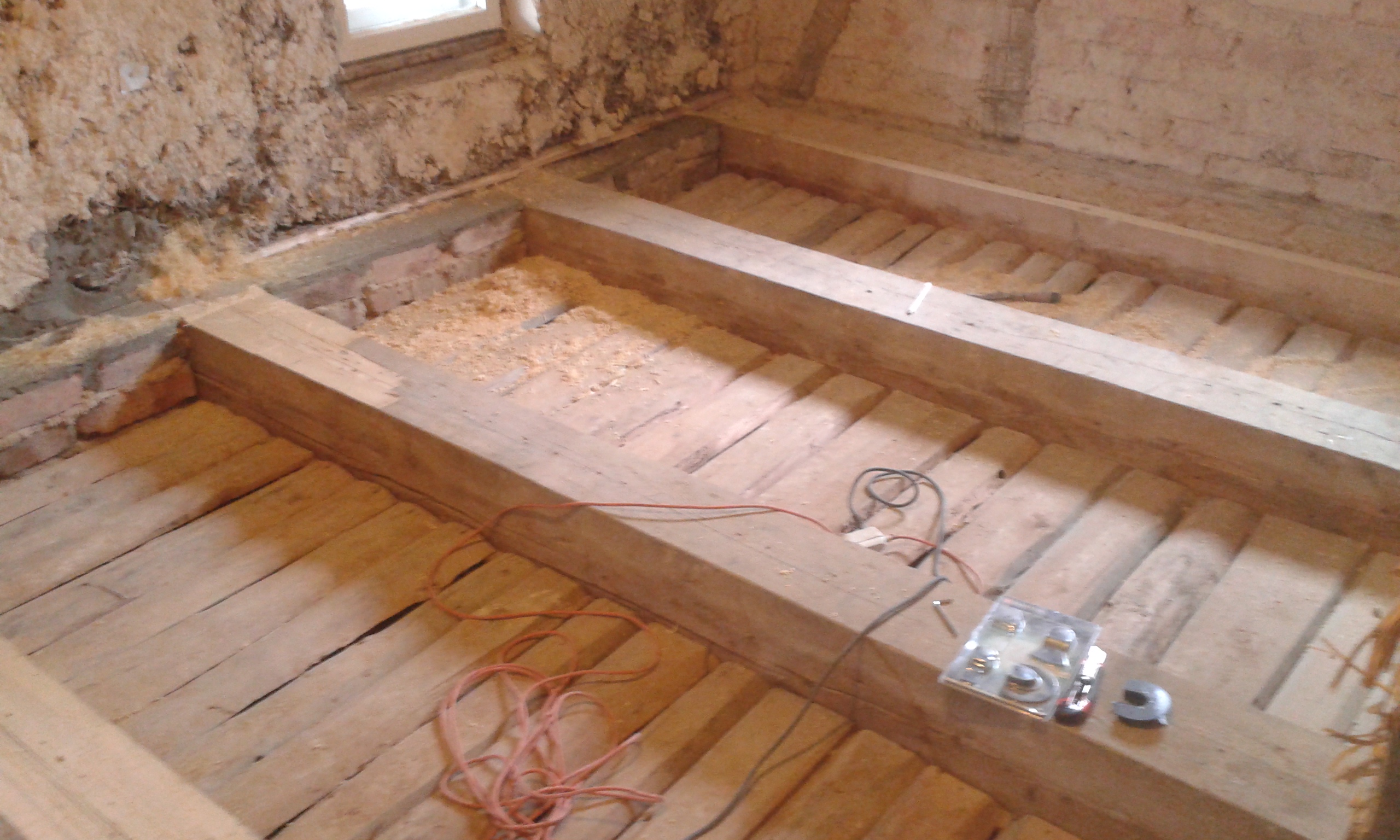 Odkryty strop drewniany z widocznymi belkami nośnymi, deskami oraz przewodami elektrycznymi podczas prac remontowych przy ścianie z oknem.