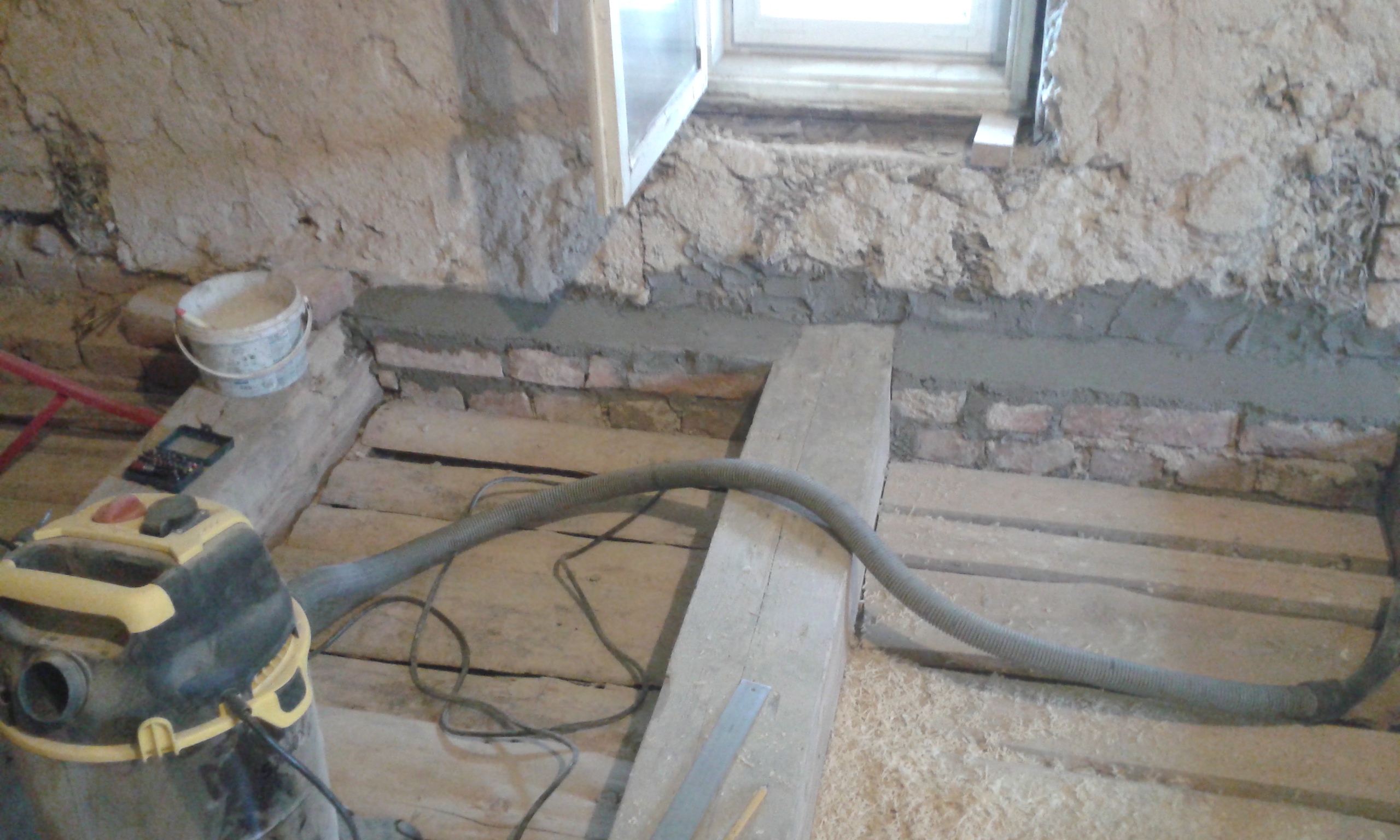 Odkryty strop drewniany przy ścianie z oknem, widoczna belka nośna, instalacja oraz sprzęt budowlany używany podczas prac remontowych.