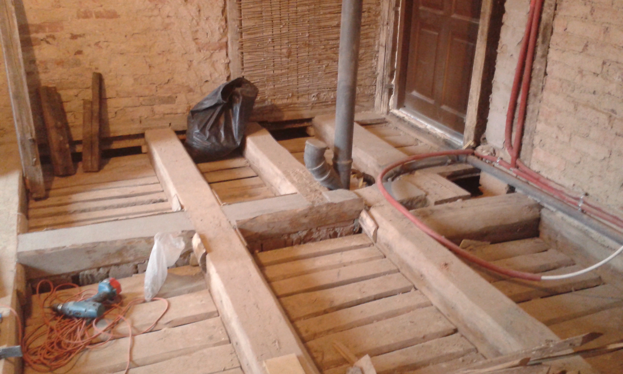 : Odkryta konstrukcja drewnianego stropu w pomieszczeniu z widoczną instalacją sanitarną, przewodami oraz narzędziami podczas prac remontowych.