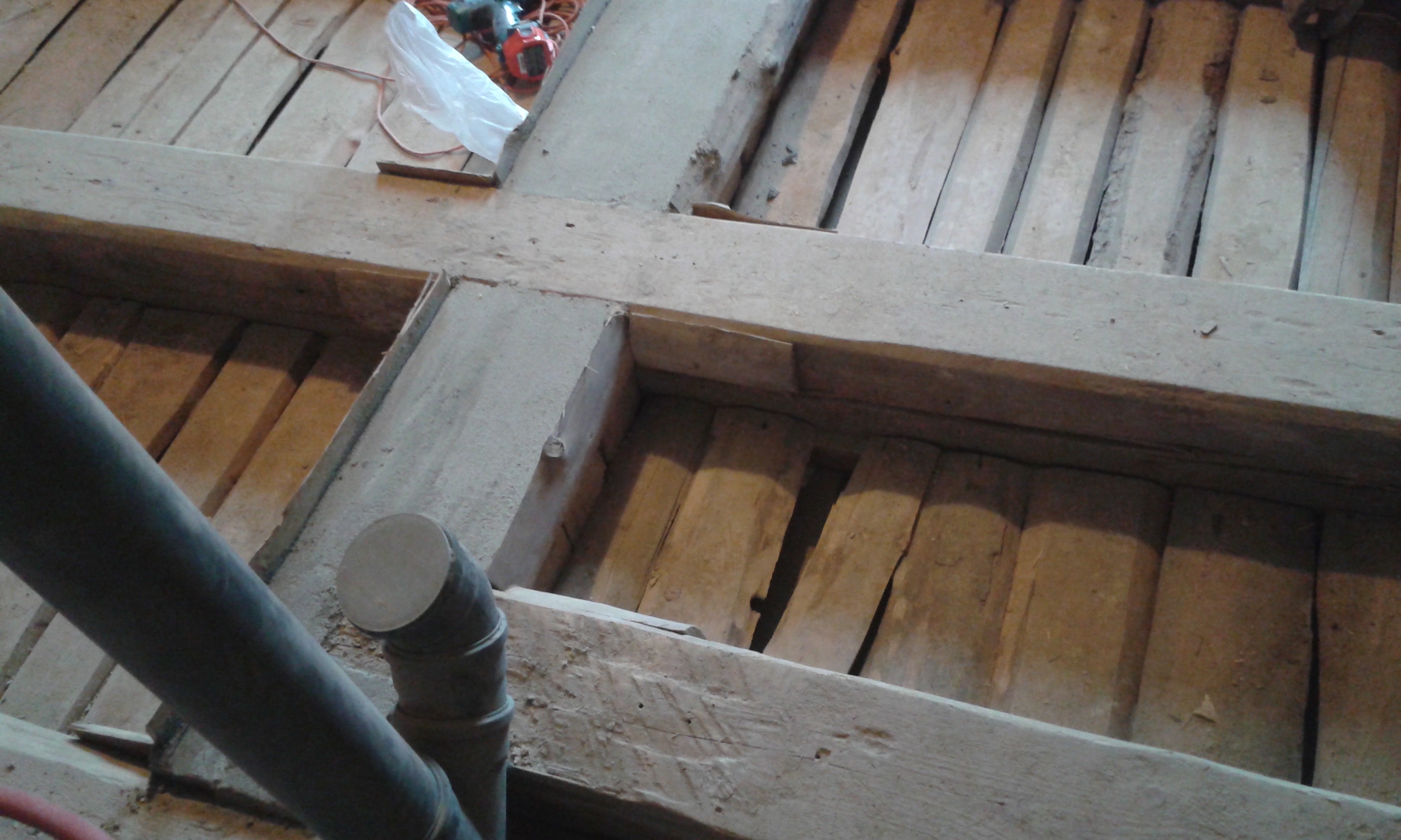 Zbliżenie na skrzyżowanie drewnianych belek stropowych z widoczną instalacją sanitarną, ukazujące konstrukcję stropu w trakcie prac remontowych.