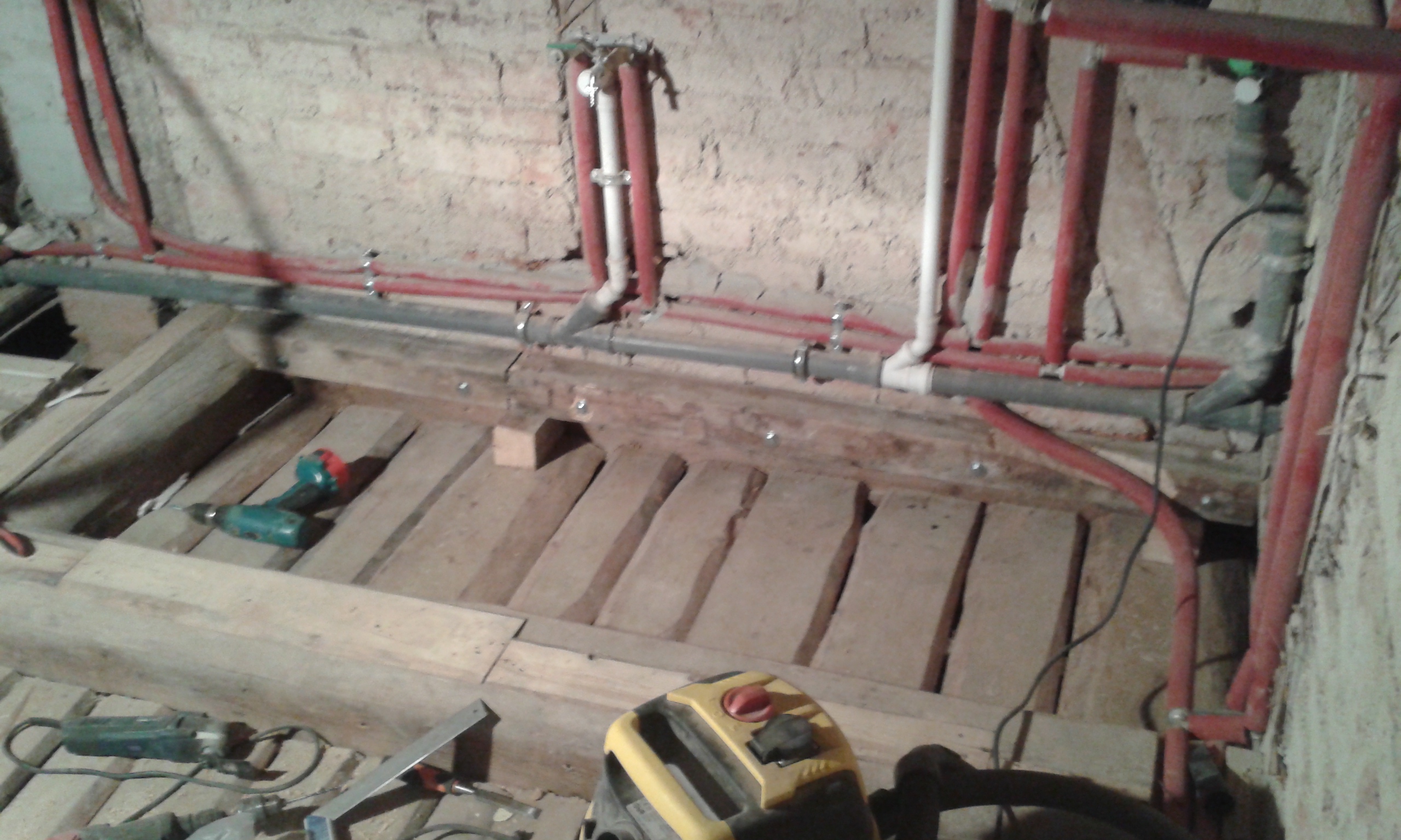 Fragment odkrytego stropu drewnianego z widocznymi instalacjami hydraulicznymi przy ścianie oraz narzędziami używanymi podczas prac remontowych.