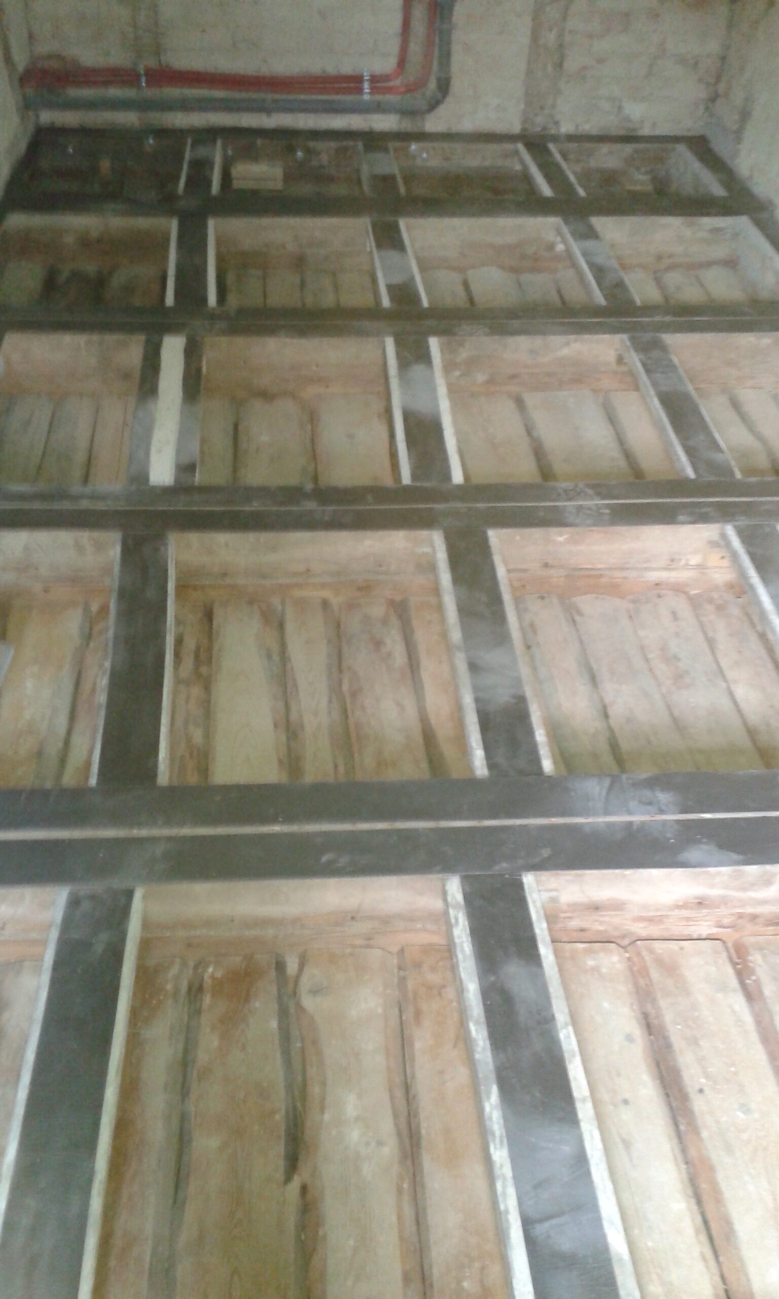 Widok na odkrytą konstrukcję stropu poddasza z widocznymi belkami nośnymi i przestrzeniami między nimi, przygotowaną do wykonania izolacji oraz dalszych prac budowlanych.