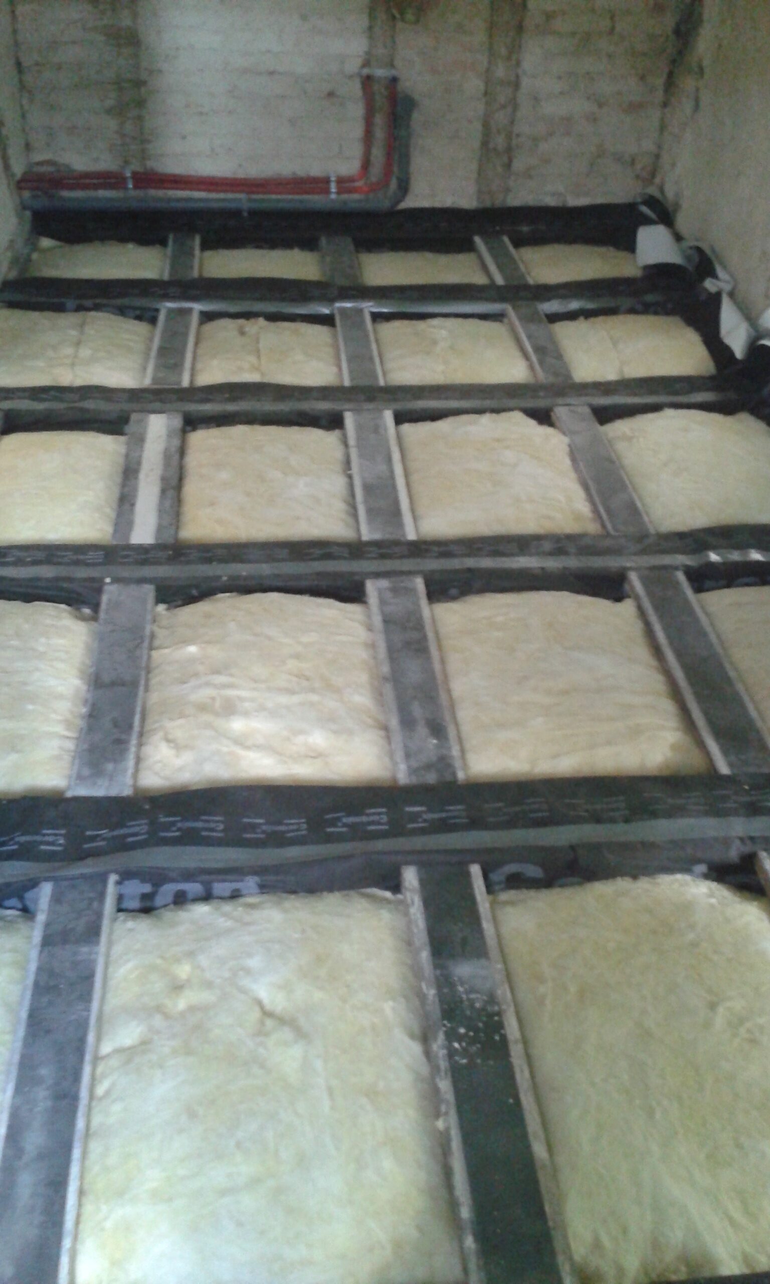 Widok na strop poddasza z zamontowaną izolacją termiczną między belkami oraz widocznymi instalacjami prowadzonymi przy ścianie.