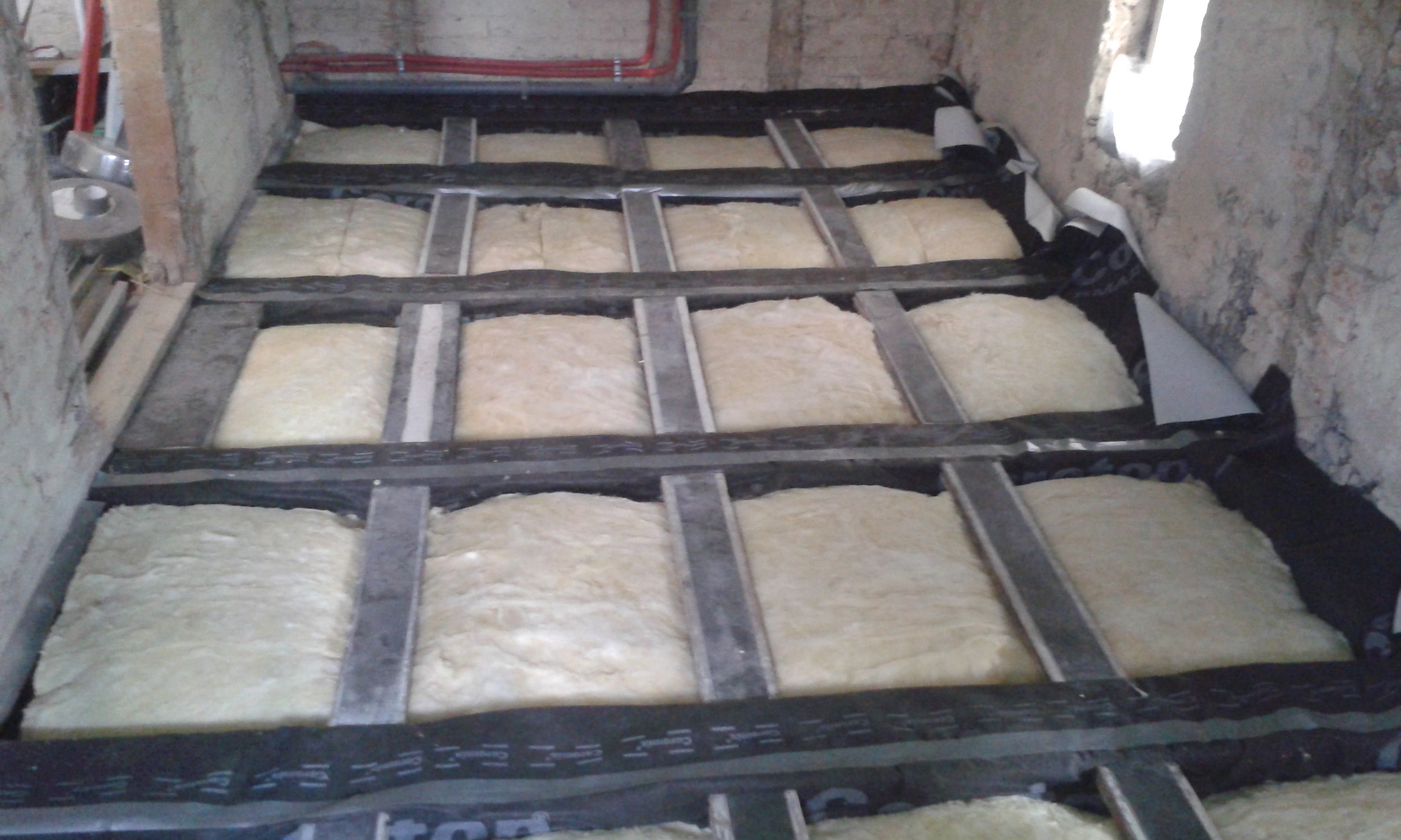 Strop poddasza z ułożoną izolacją termiczną pomiędzy belkami konstrukcyjnymi, zabezpieczoną warstwą techniczną i przygotowaną do montażu podłogi.