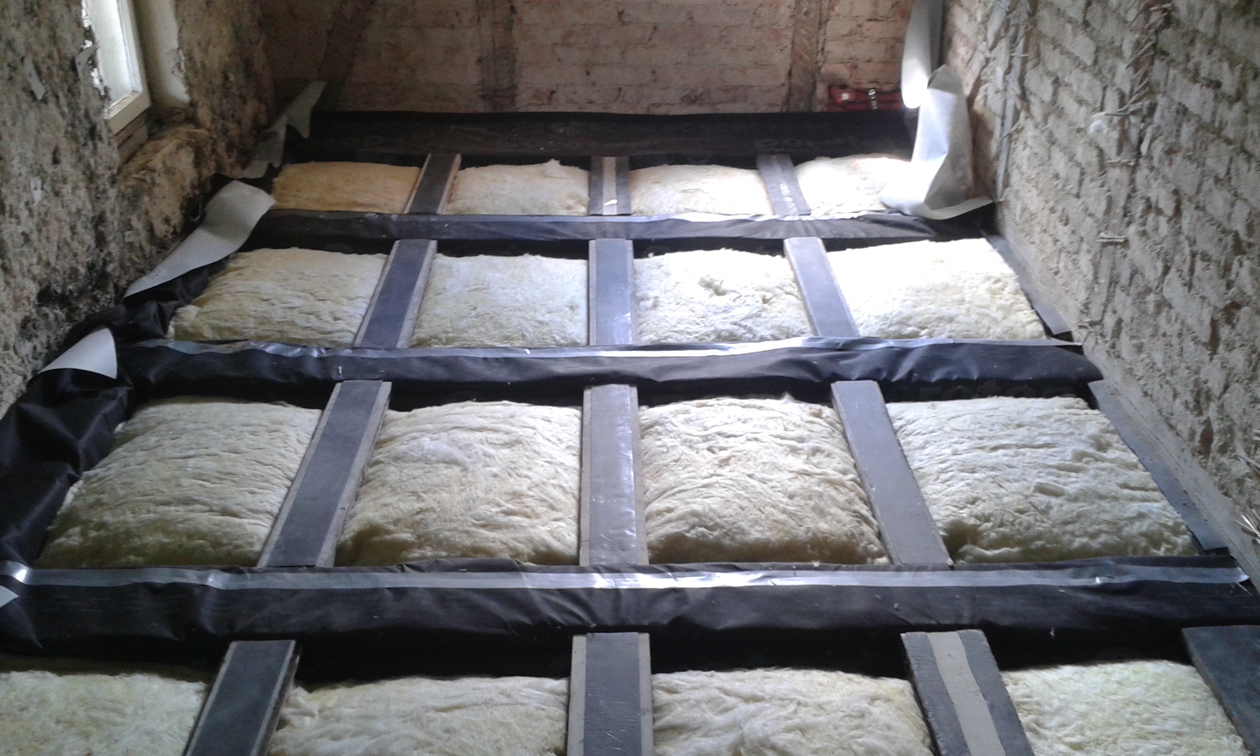 Widok na strop poddasza z ułożoną izolacją termiczną między belkami, zabezpieczoną taśmami montażowymi, przygotowany do dalszych prac wykończeniowych.