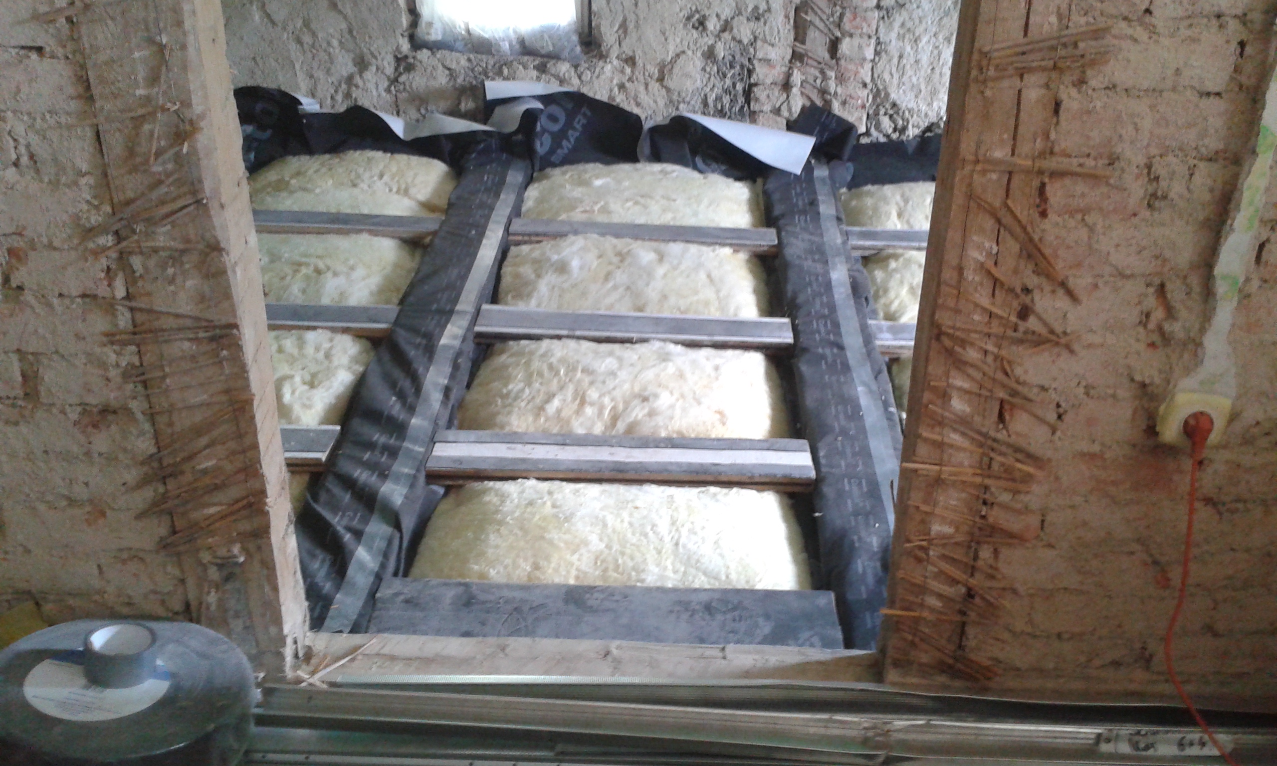 Widok na konstrukcję stropu z ułożoną izolacją termiczną między belkami, widoczne taśmy montażowe oraz surowe mury w trakcie prac budowlanych.