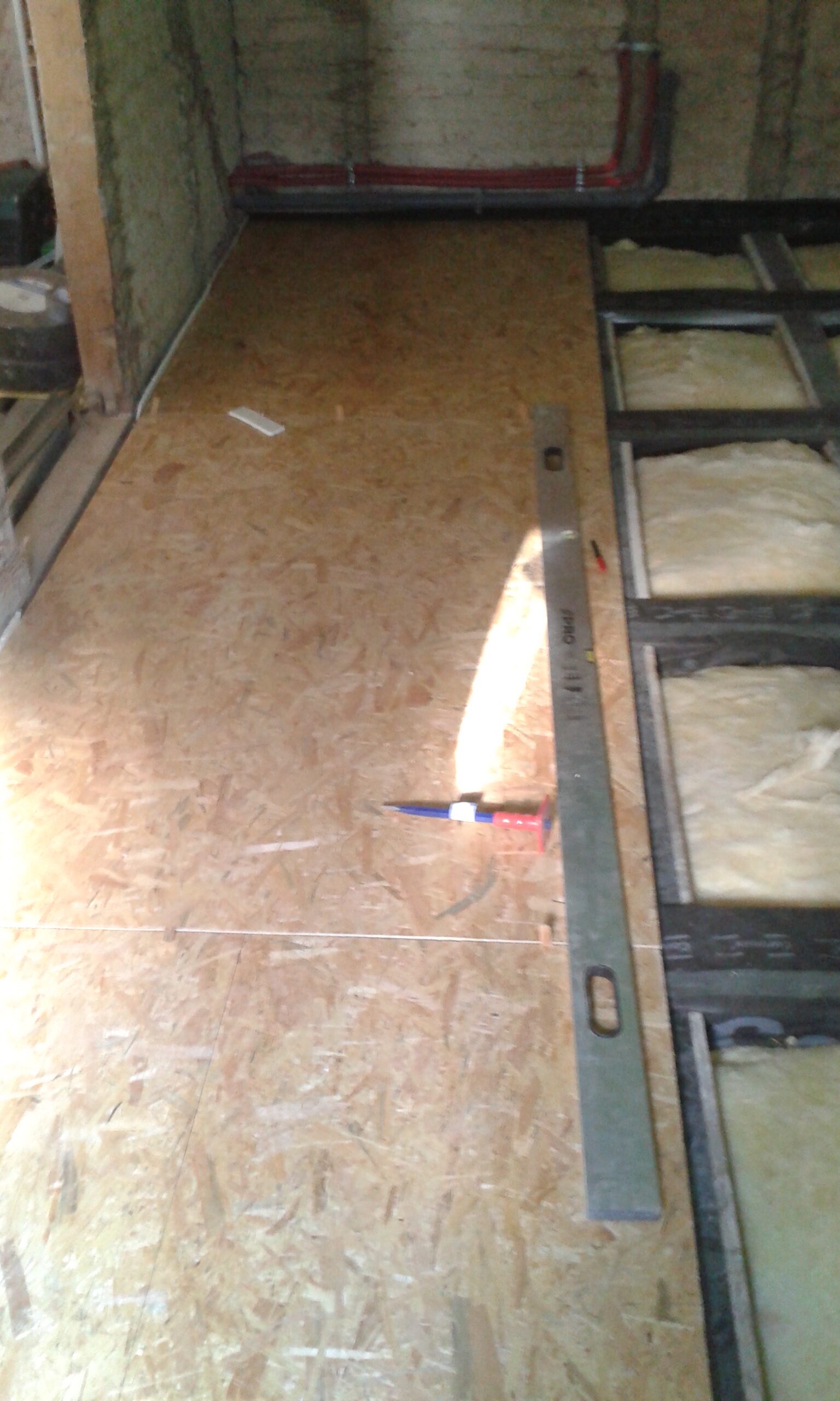 Etap montażu podłogi z płyt OSB na poddaszu, widoczne narzędzia pomiarowe, belki stropowe z izolacją oraz instalacje prowadzone przy ścianie.