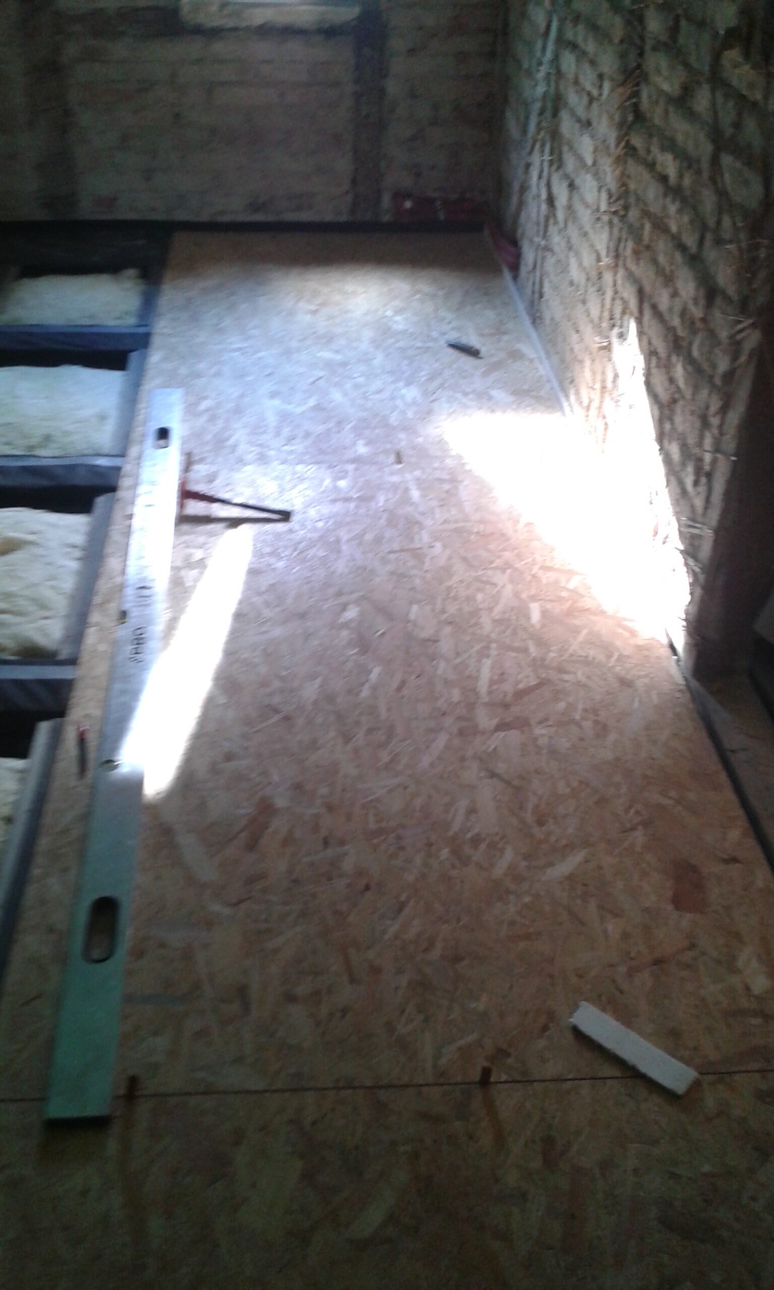 Fragment poddasza z układaną podłogą z płyt OSB, widocznymi belkami stropowymi, narzędziami oraz naturalnym światłem wpadającym przez okno.