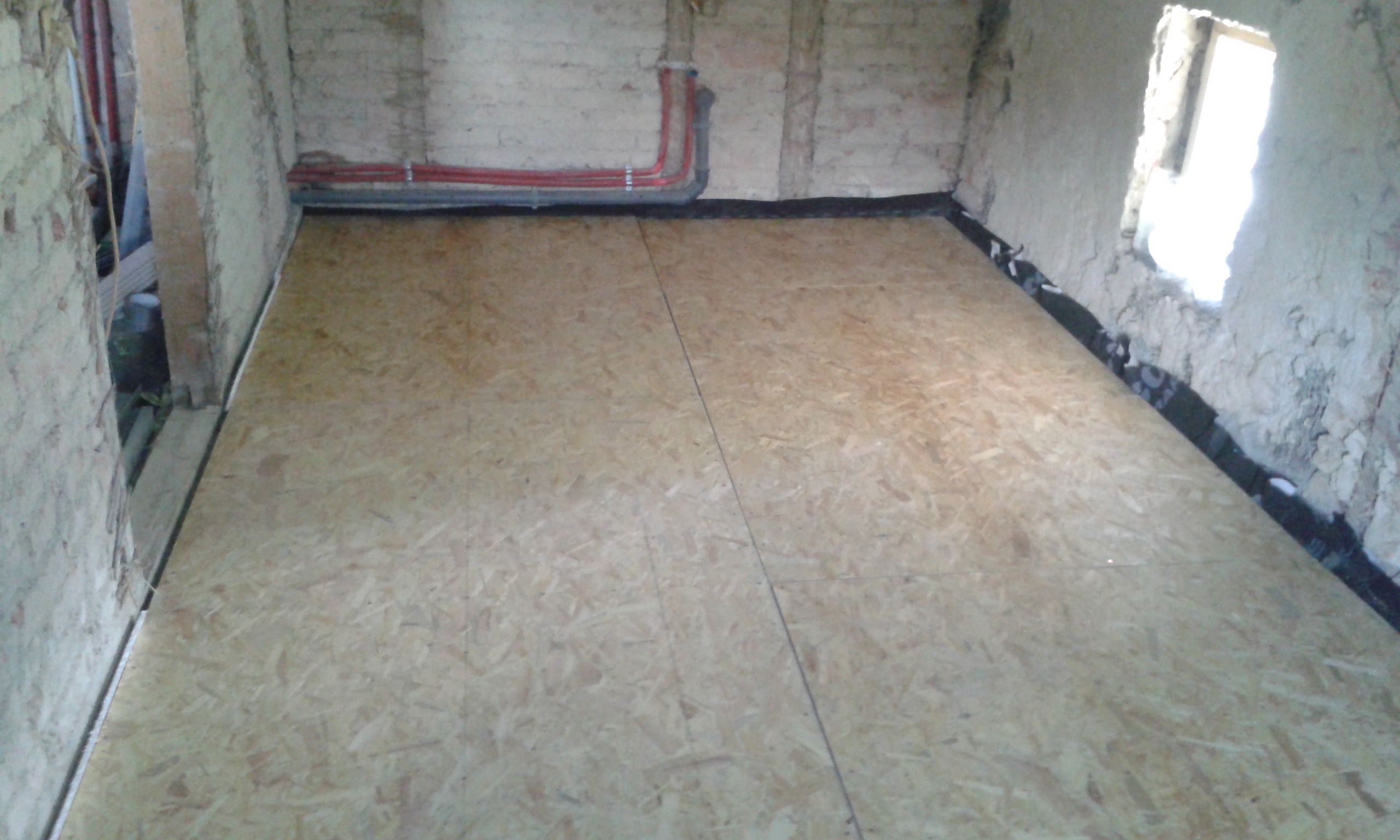 Pomieszczenie w trakcie prac budowlanych z ułożoną podłogą z płyt OSB, widoczną instalacją przy tylnej ścianie oraz doświetleniem z bocznego okna.