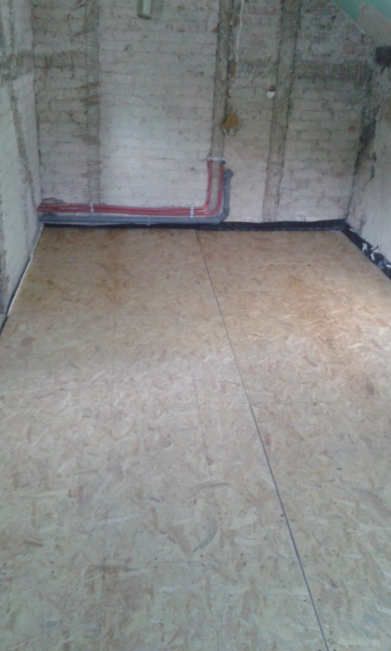 Wnętrze pomieszczenia z ułożoną podłogą z płyt OSB oraz widoczną instalacją prowadzoną przy ścianie z surowej cegły.