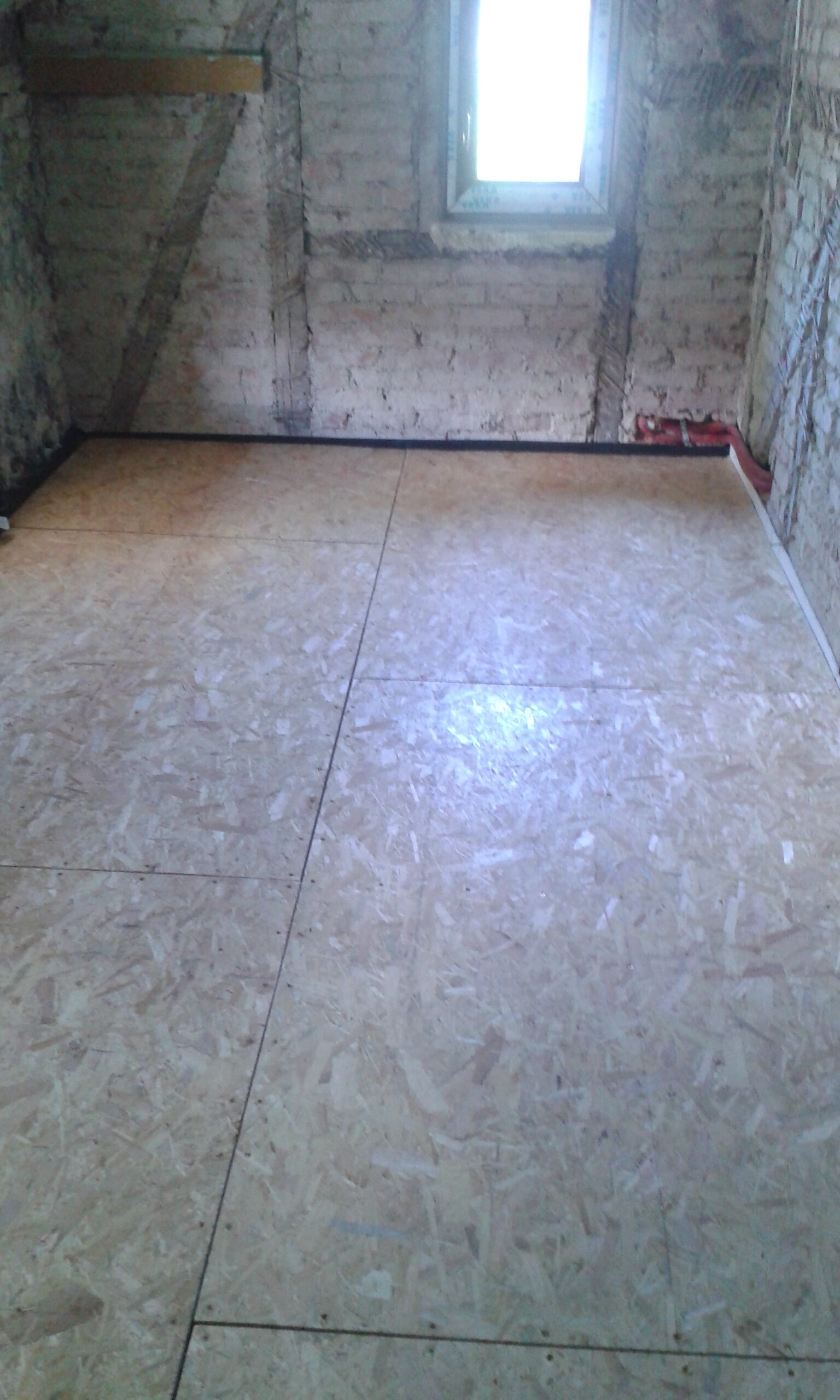 Pomieszczenie na poddaszu z ułożoną podłogą z płyt OSB, surową ceglaną ścianą oraz oknem doświetlającym wnętrze od frontu.