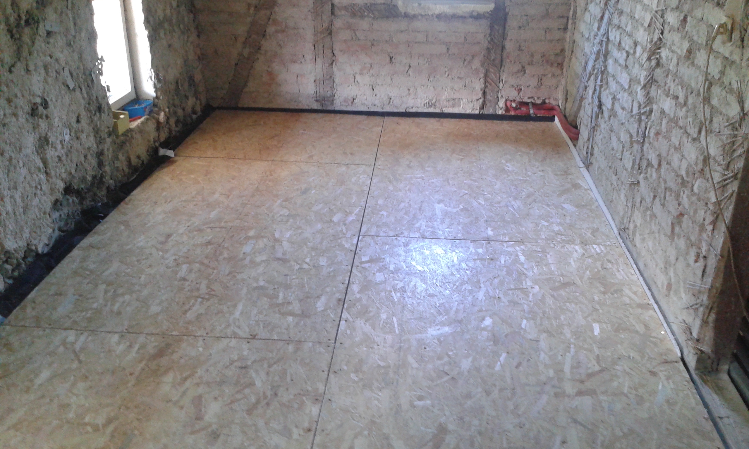 Poddasze z równo ułożonymi płytami OSB tworzącymi stabilną podłogę, widoczne surowe ściany z cegły oraz instalacje prowadzone przy krawędziach.