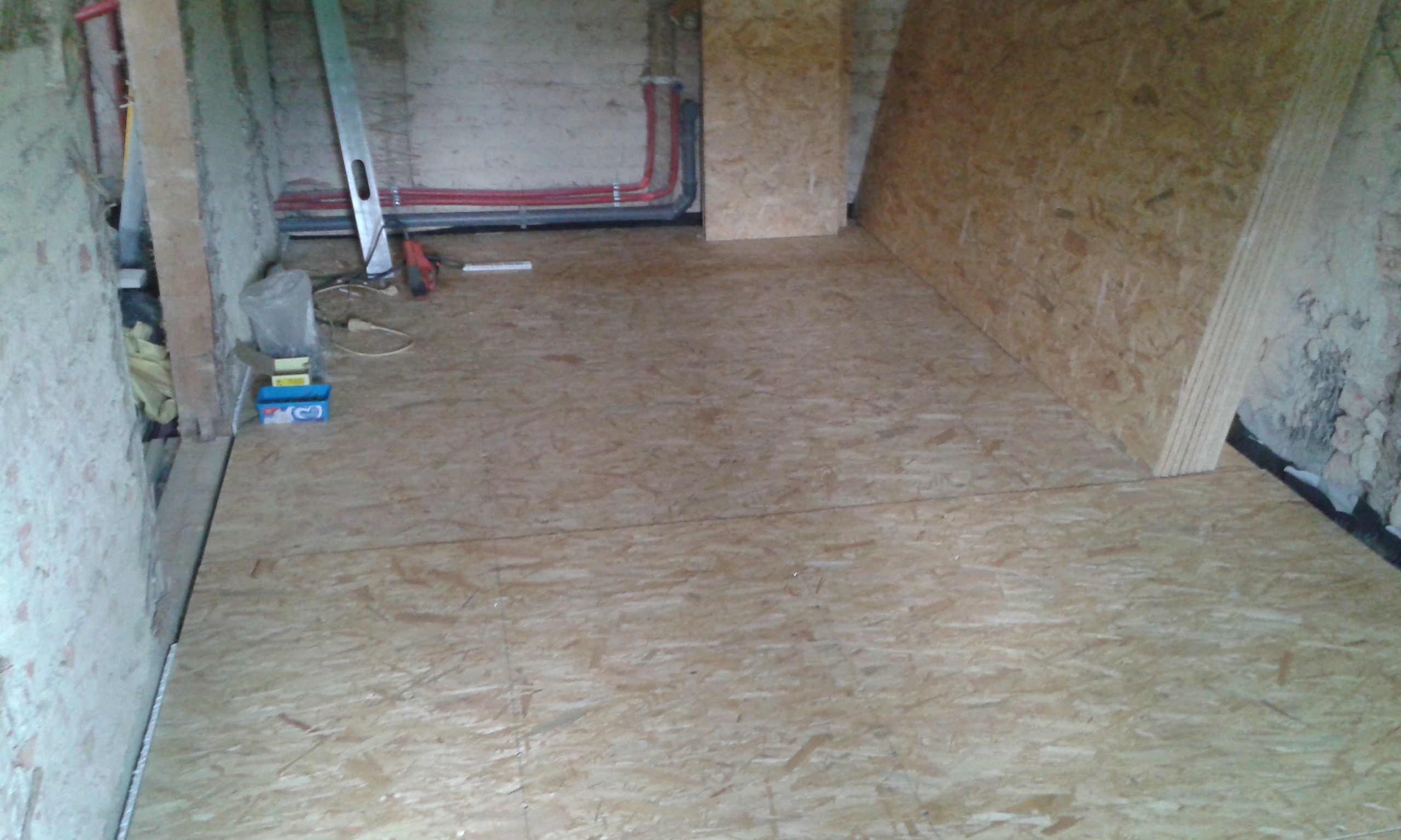 Pomieszczenie w trakcie prac budowlanych z ułożoną podłogą z płyt OSB, widocznymi instalacjami przy ścianach oraz przygotowaniem do dalszych etapów wykończenia.