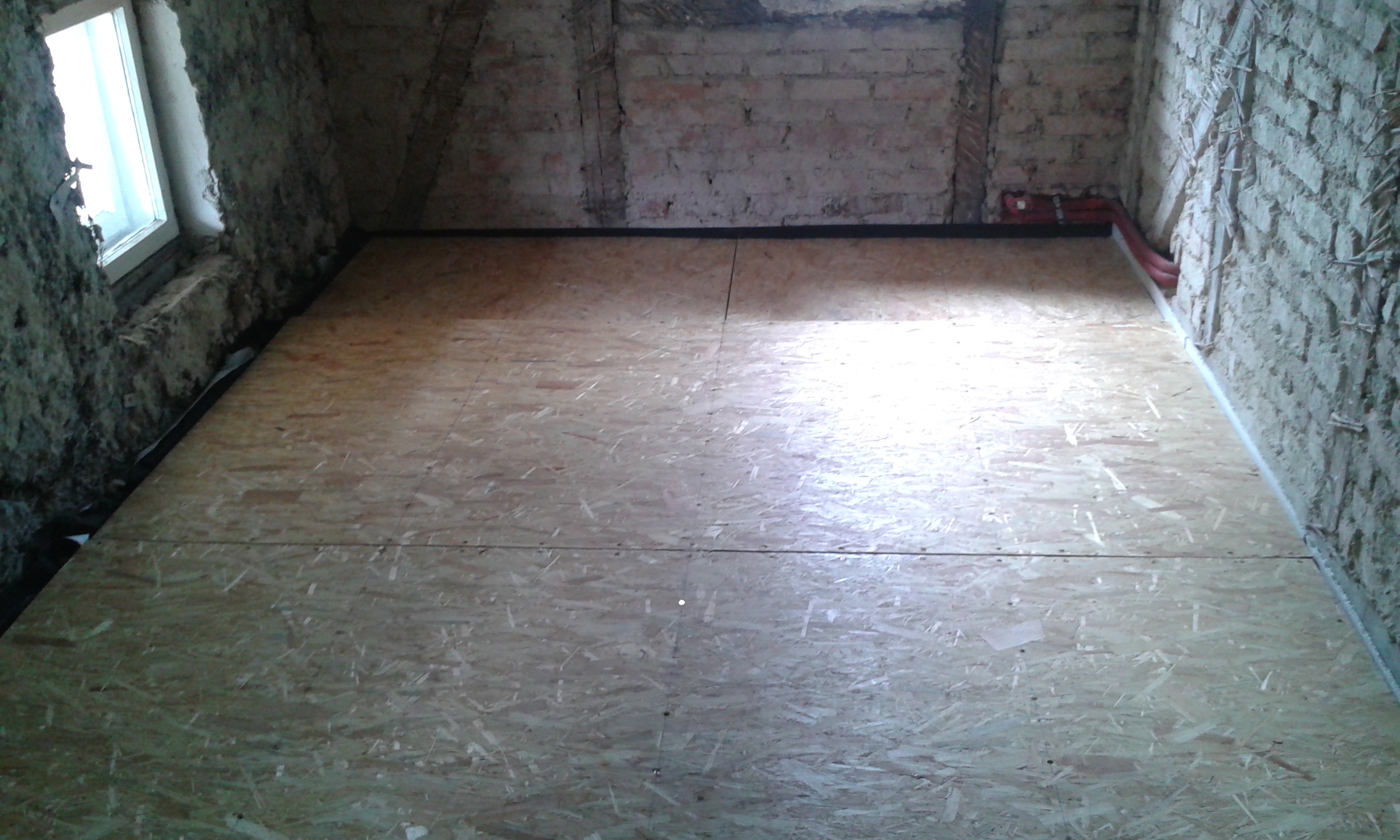 Pomieszczenie na poddaszu z równomiernie ułożoną podłogą z płyt OSB, surowymi cegłami na ścianach oraz widocznym doświetleniem z okna.