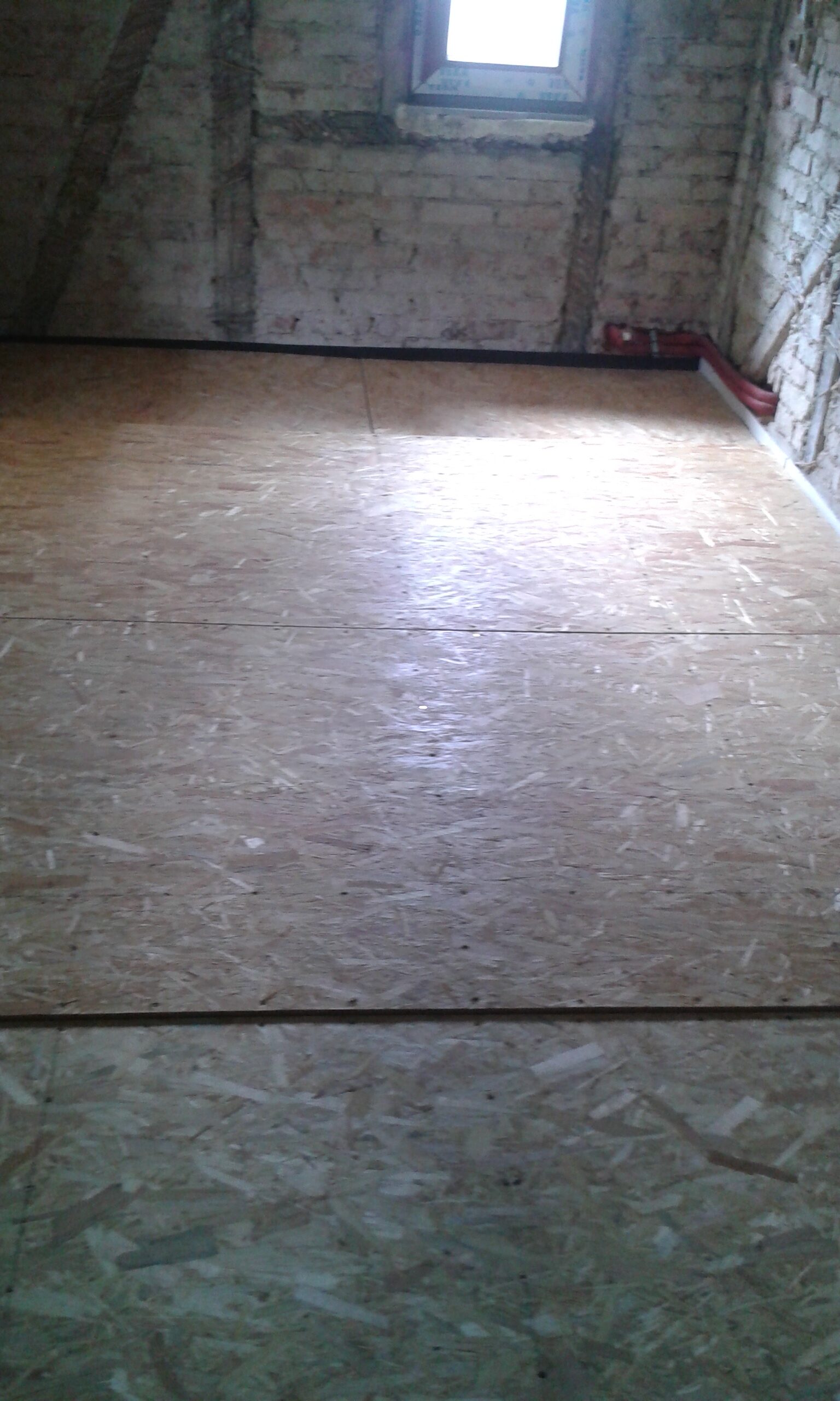 Poddasze w trakcie prac budowlanych z ułożoną podłogą z płyt OSB, surowymi ścianami z cegły oraz niewielkim oknem doświetlającym wnętrze.