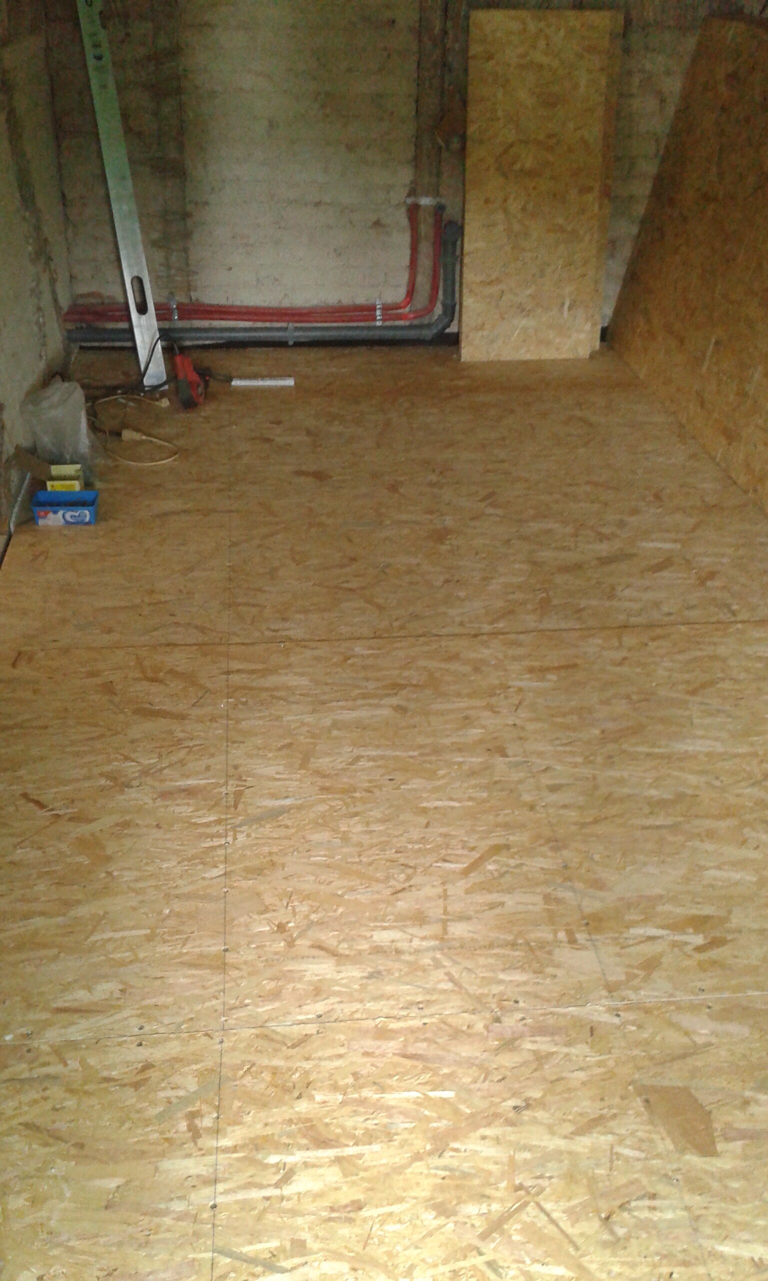 Wnętrze pomieszczenia w trakcie prac budowlanych z wykonaną podłogą z płyt OSB, widocznymi instalacjami przy ścianie oraz narzędziami roboczymi.