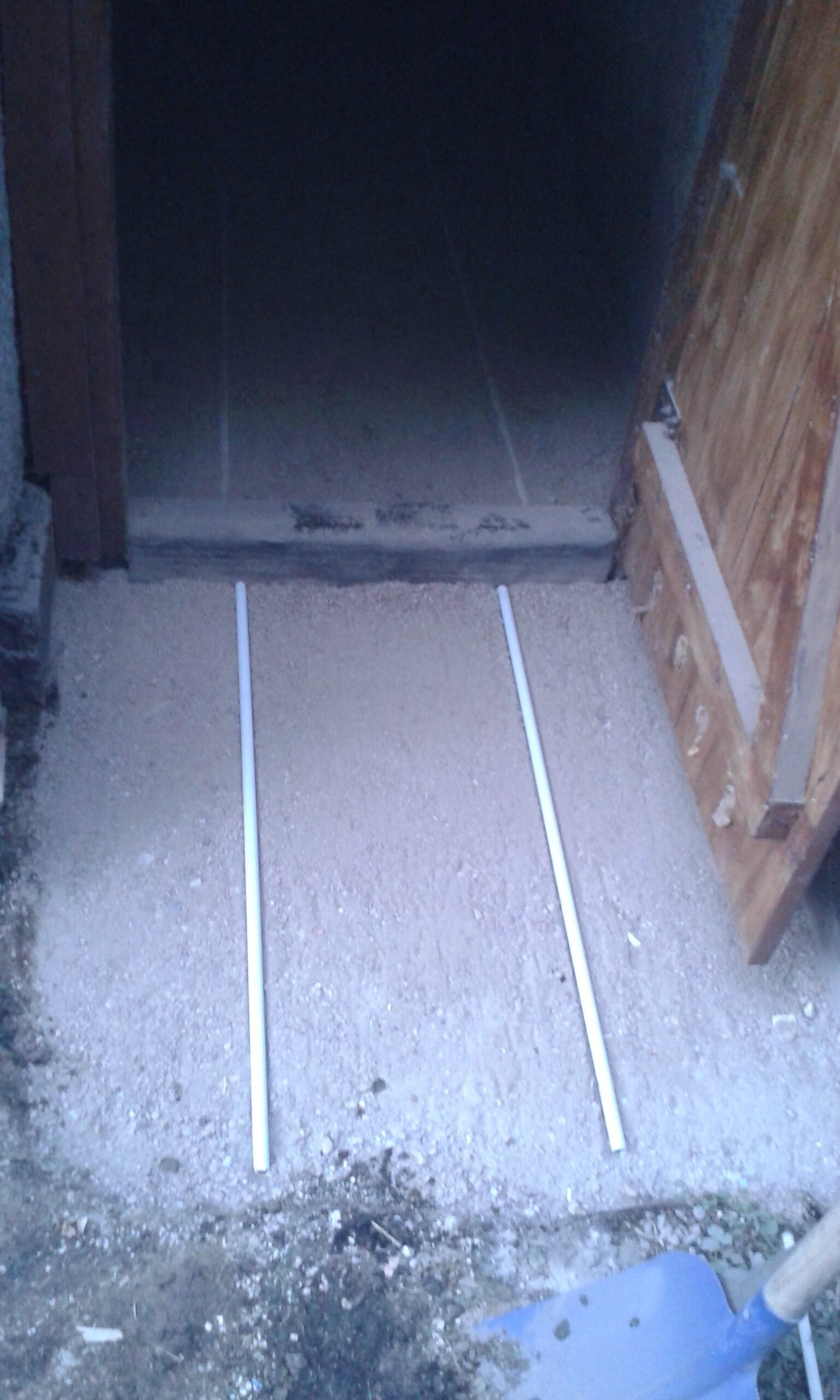 Wyrównane podłoże przy wejściu do pomieszczenia, przygotowane pod ułożenie kostki brukowej, z widocznymi listwami prowadzącymi i surowym progiem.