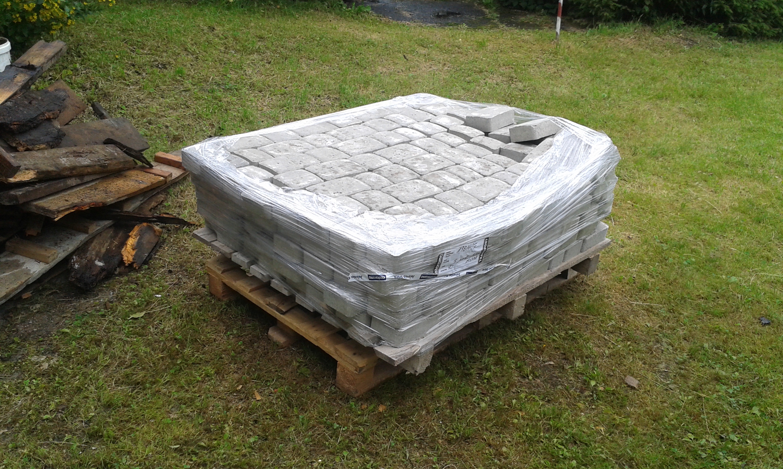 Zabezpieczona folią paleta z kostką brukową ułożoną na drewnianym podeście, przygotowana do wykorzystania podczas prac przy nawierzchni.