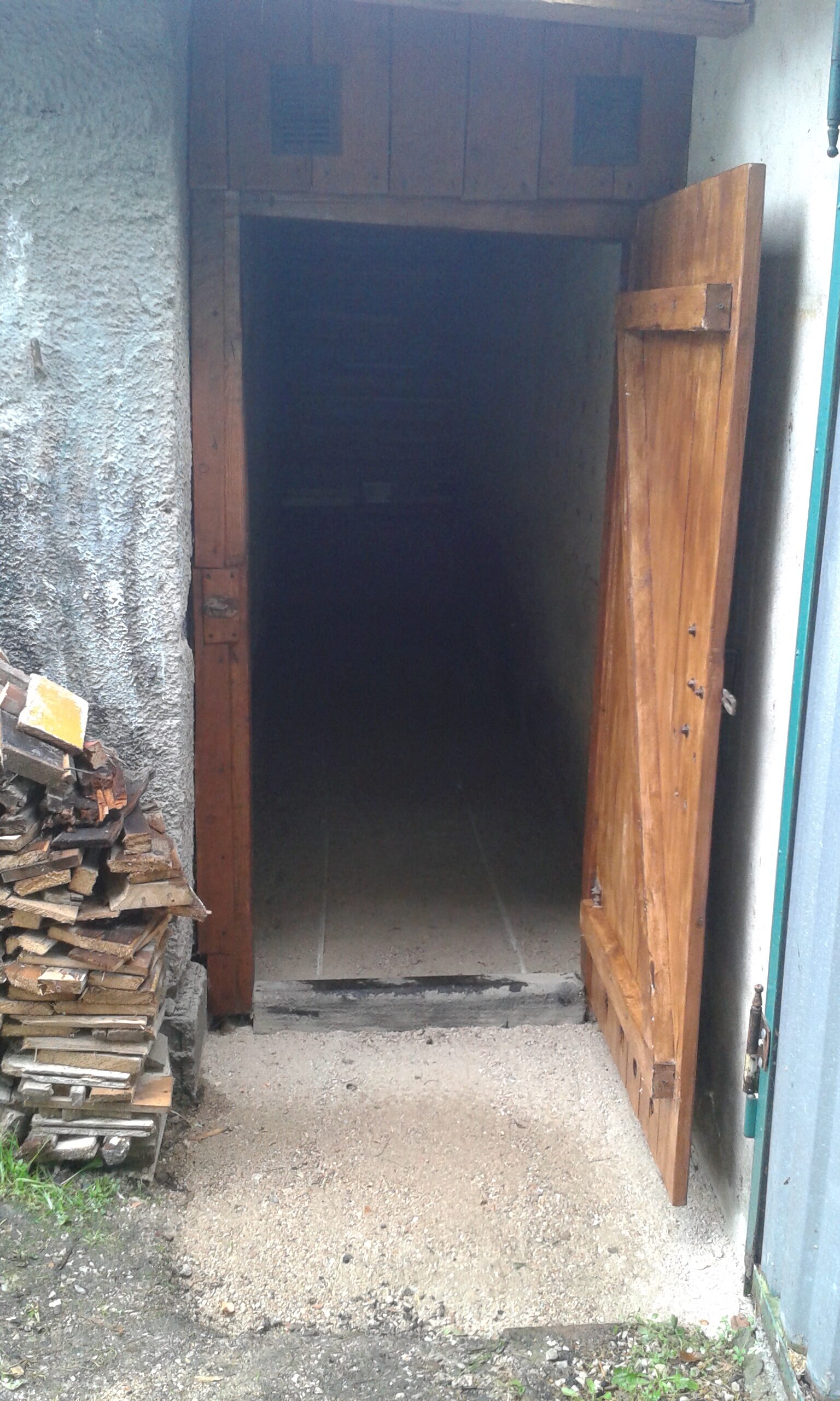 Wejście do pomieszczenia przed rozpoczęciem prac brukarskich, z widocznym nieutwardzonym podłożem, drewnianymi drzwiami i surowym progiem.