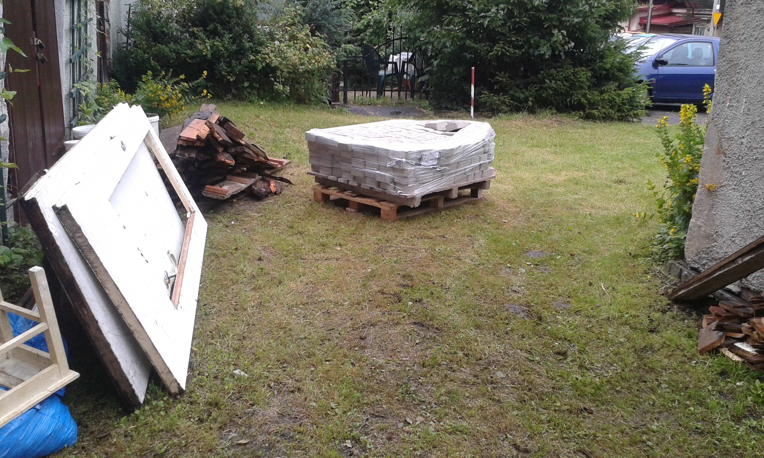 Paleta z kostką brukową przygotowaną do prac, ustawiona na trawniku obok budynku, z widocznymi materiałami i elementami zaplecza robót.