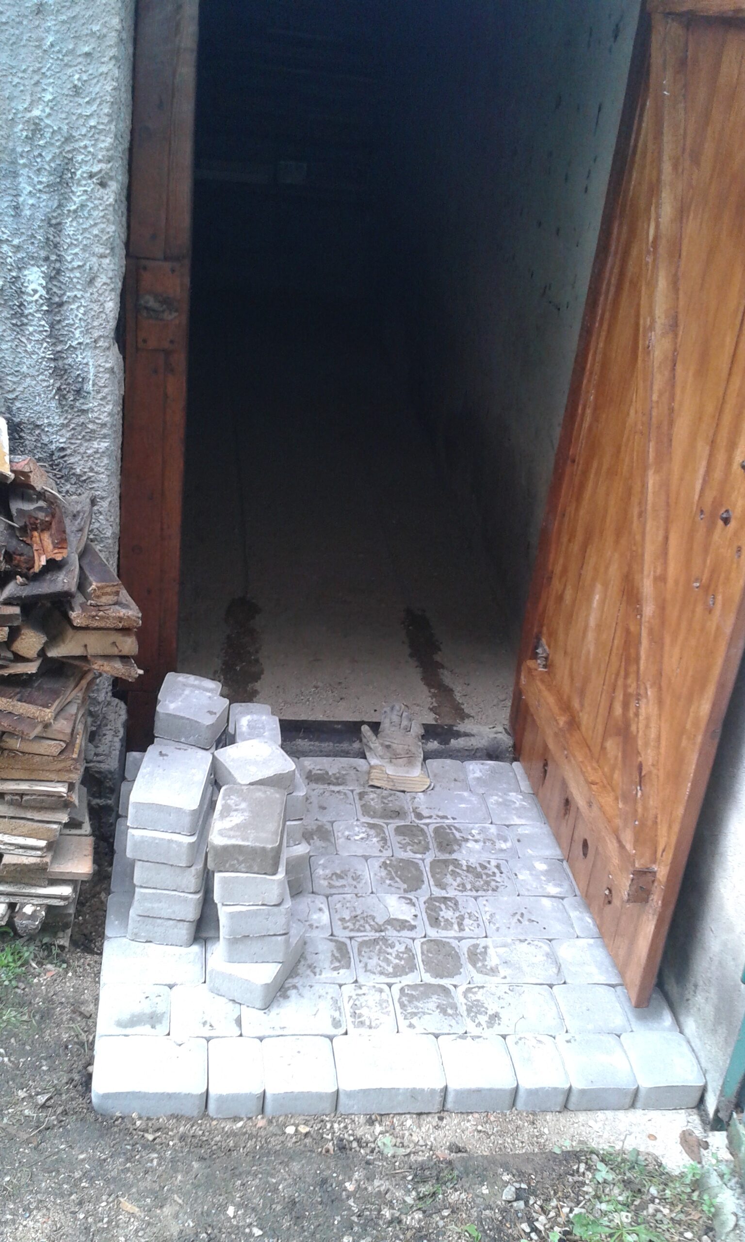 Wejście do pomieszczenia w trakcie prac brukarskich, z częściowo ułożoną kostką brukową, przygotowanym progiem i zapasem materiału obok drzwi.