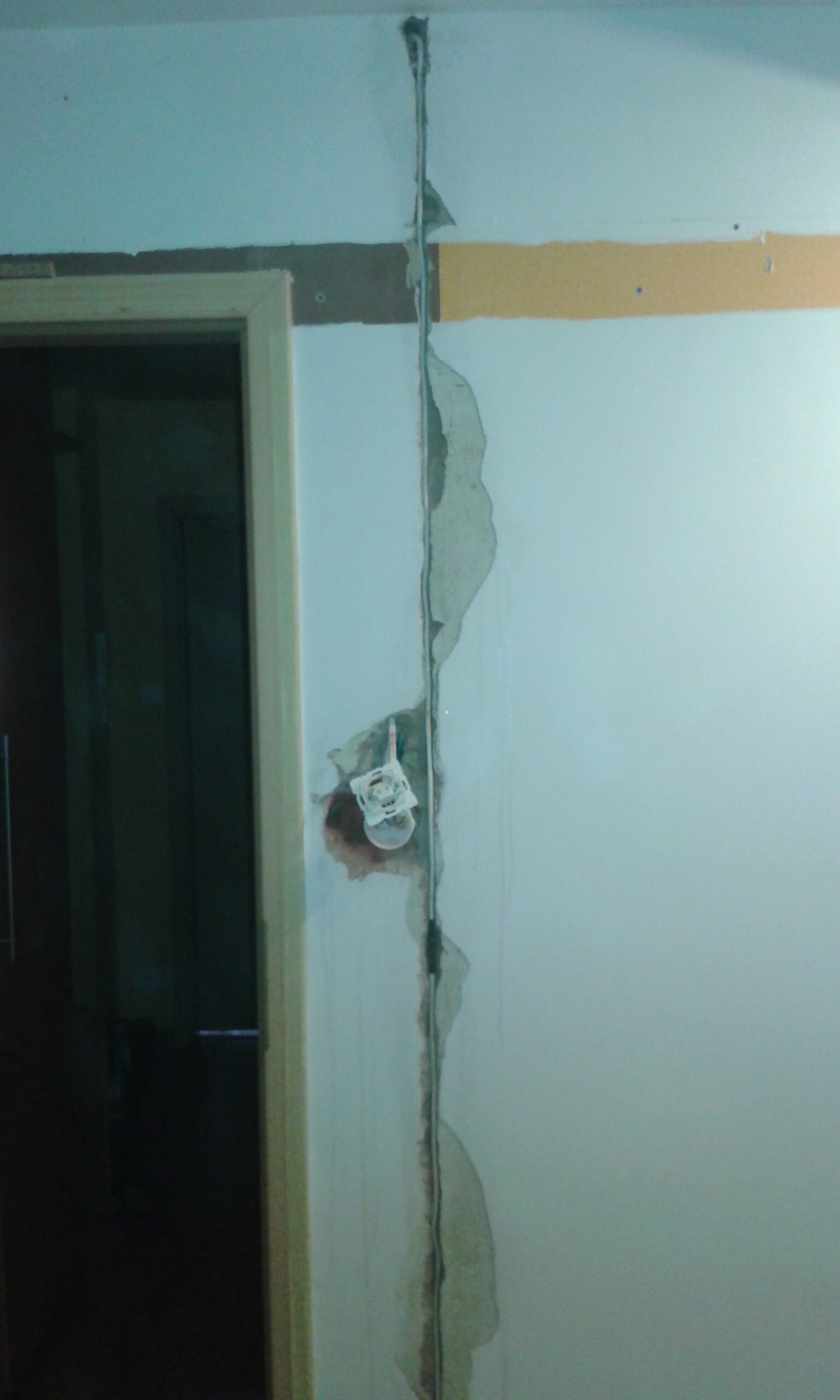 Pionowa bruzda instalacyjna w ścianie obok drzwi z odsłoniętym przewodem i puszką elektryczną podczas prac remontowych.