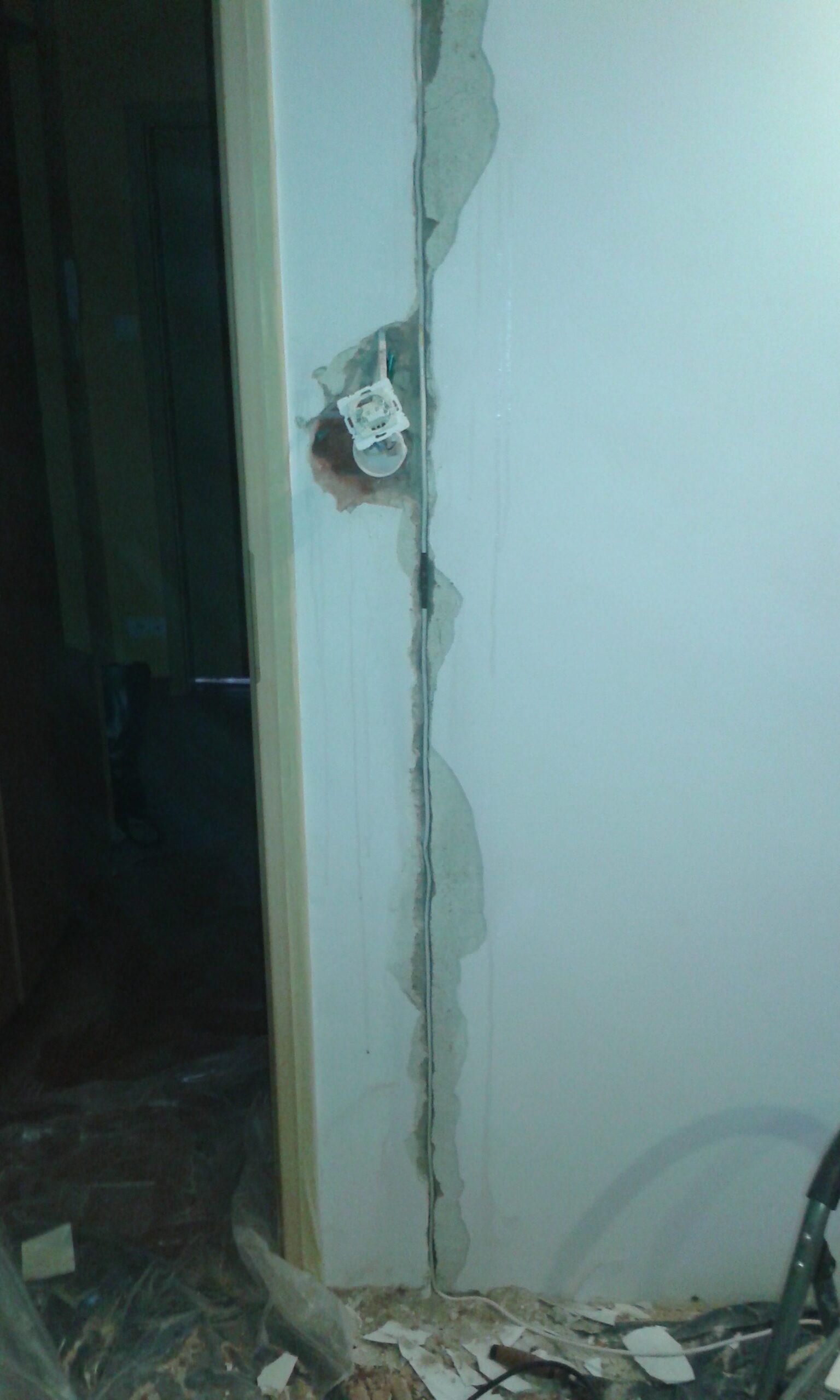 Pionowa bruzda w ścianie obok ościeżnicy drzwi z odsłoniętą puszką elektryczną i fragmentami tynku w trakcie prac remontowych.