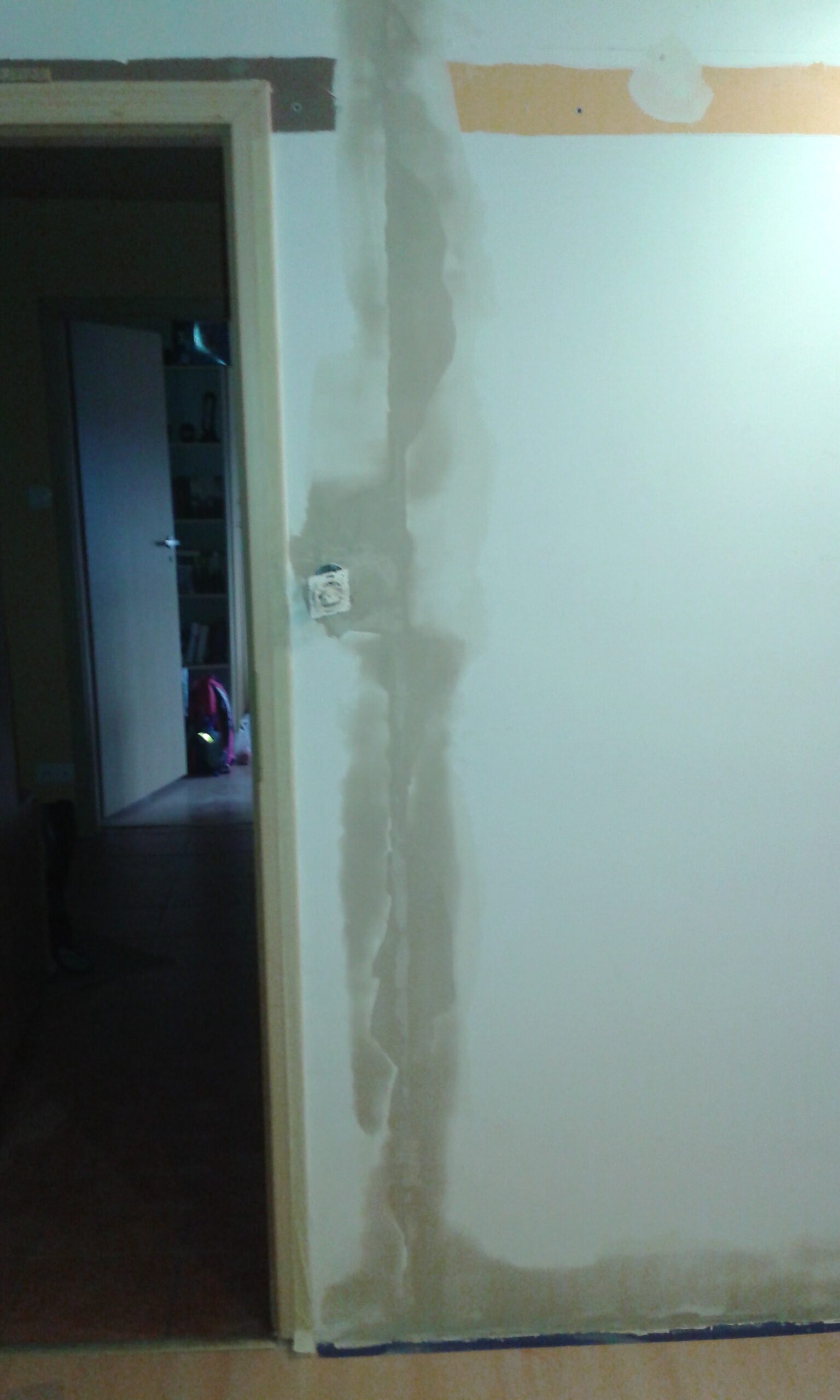 Ściana w trakcie remontu z pionową bruzdą po instalacji elektrycznej, częściowo zaszpachlowaną, z widocznym gniazdkiem obok ościeżnicy drzwi.