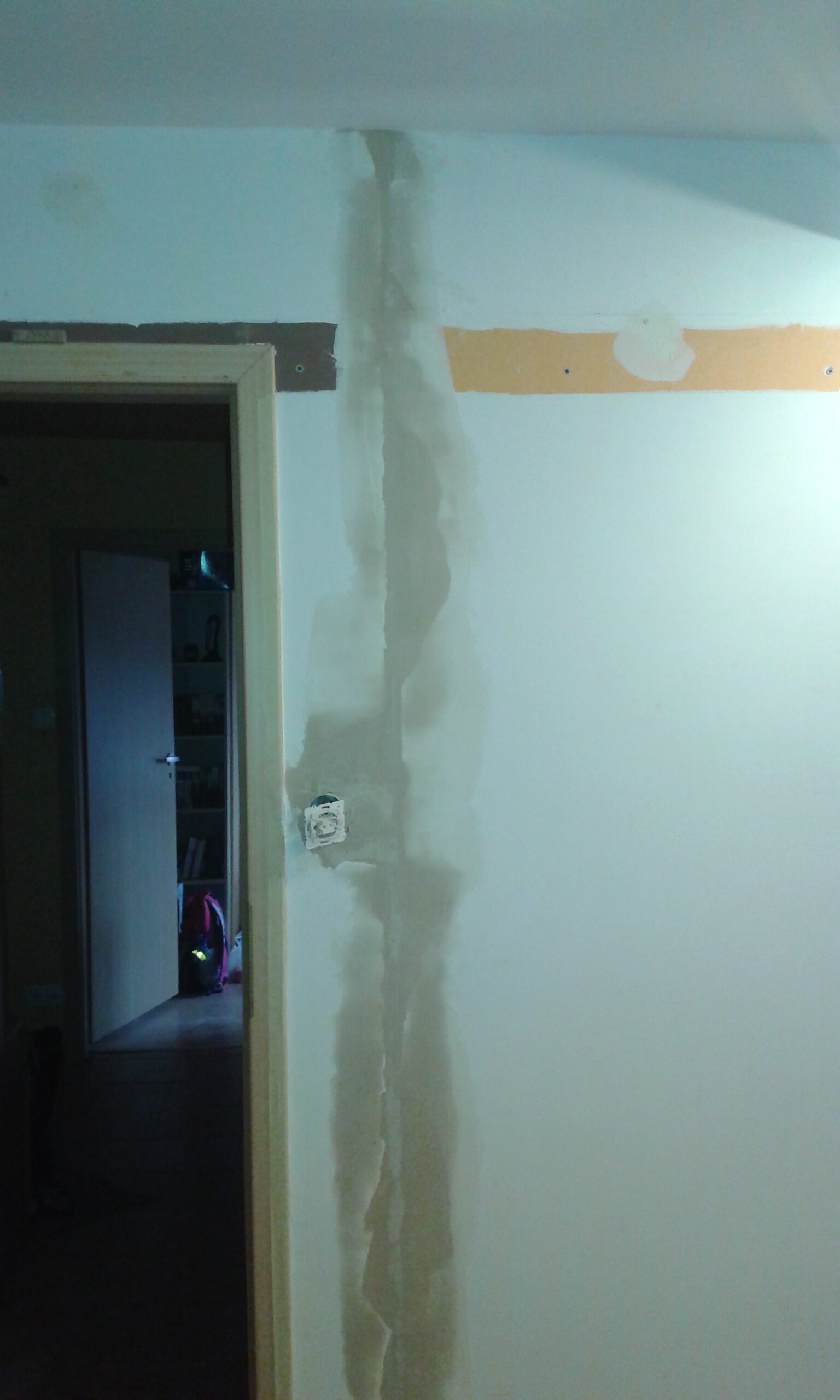 Ściana w trakcie remontu z pionową, zaszpachlowaną bruzdą po instalacji elektrycznej oraz widocznym gniazdkiem przy ościeżnicy drzwi.