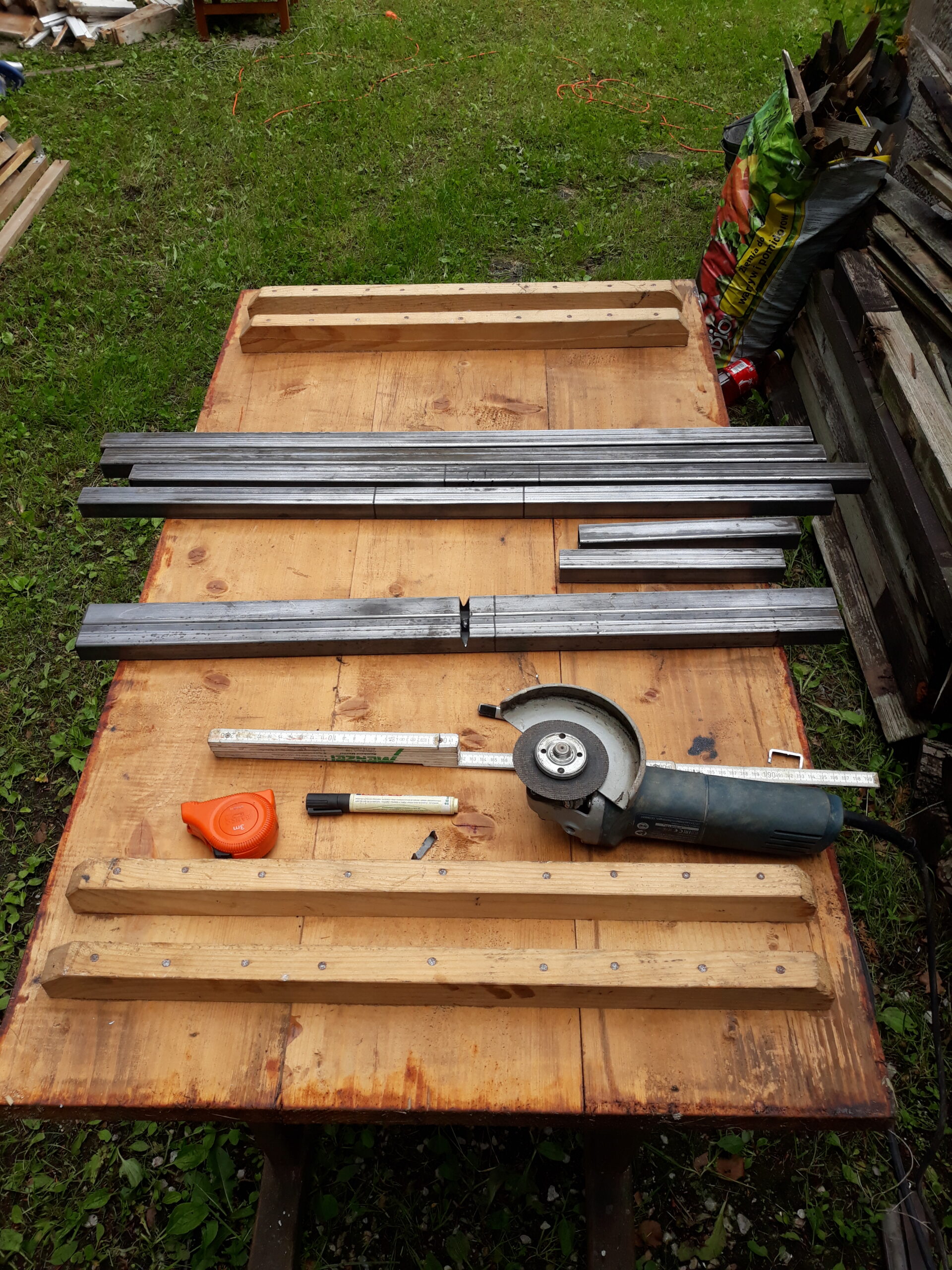 Metalowe profile, drewniane belki i narzędzia ułożone na stole roboczym, przygotowane do wykonania konstrukcji ławki ogrodowej.