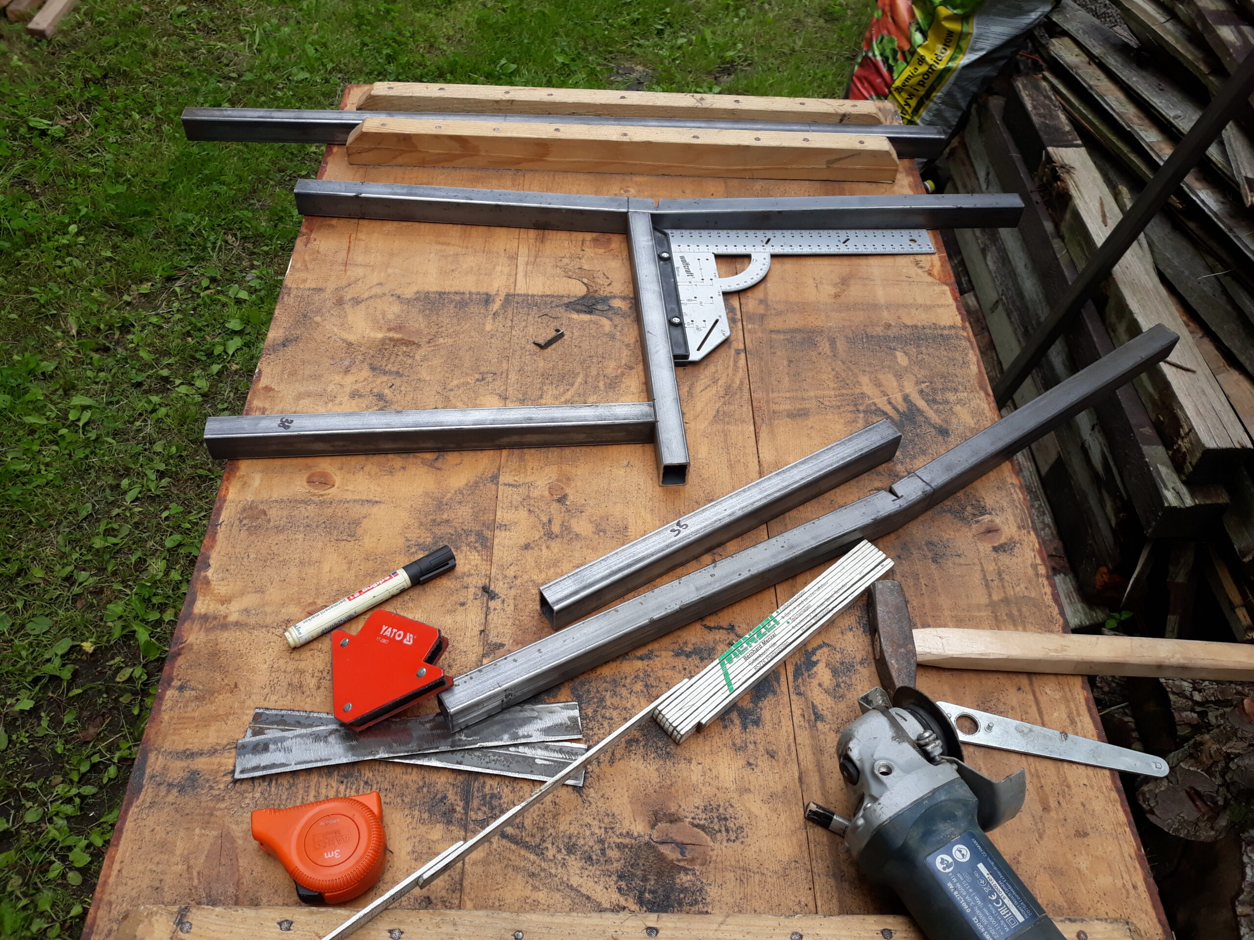 Metalowe profile i narzędzia ułożone na stole roboczym, pokazujące etap przygotowania elementów ramy ławki do spawania i montażu.