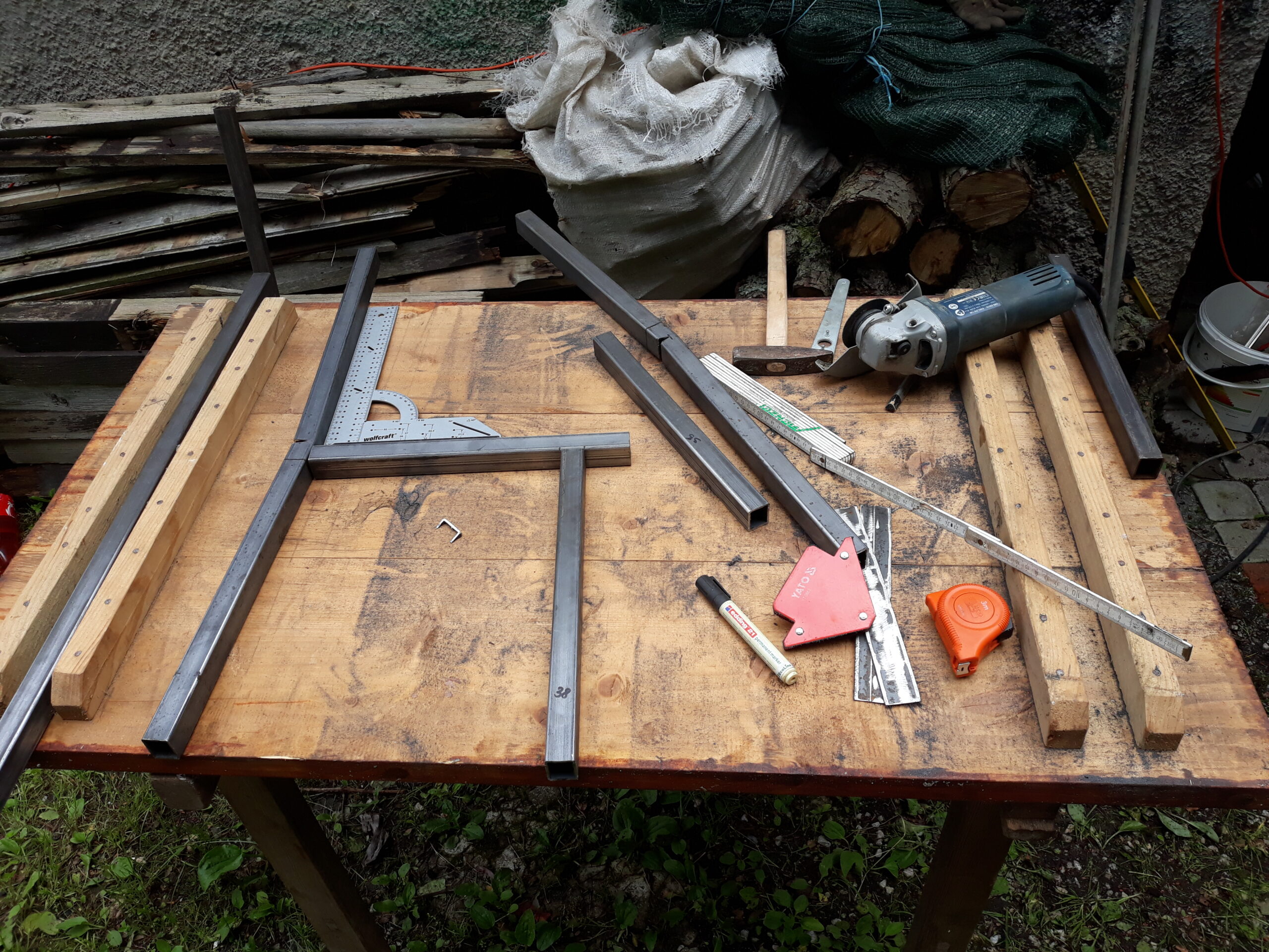 Stół roboczy z metalowymi profilami, narzędziami pomiarowymi i szlifierką kątową, przygotowany do prac spawalniczych przy konstrukcji ławki.