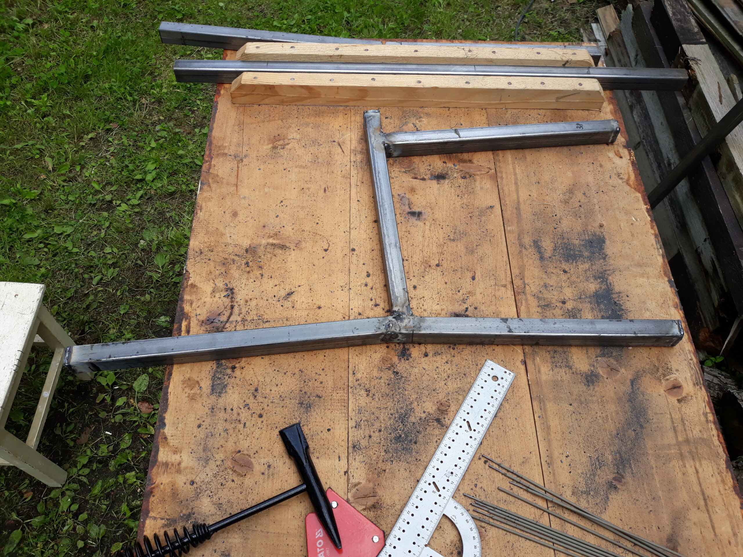 Metalowe elementy ramy ławki ułożone na stole roboczym, z widocznymi narzędziami pomiarowymi i przygotowaniem do dalszych prac spawalniczych.