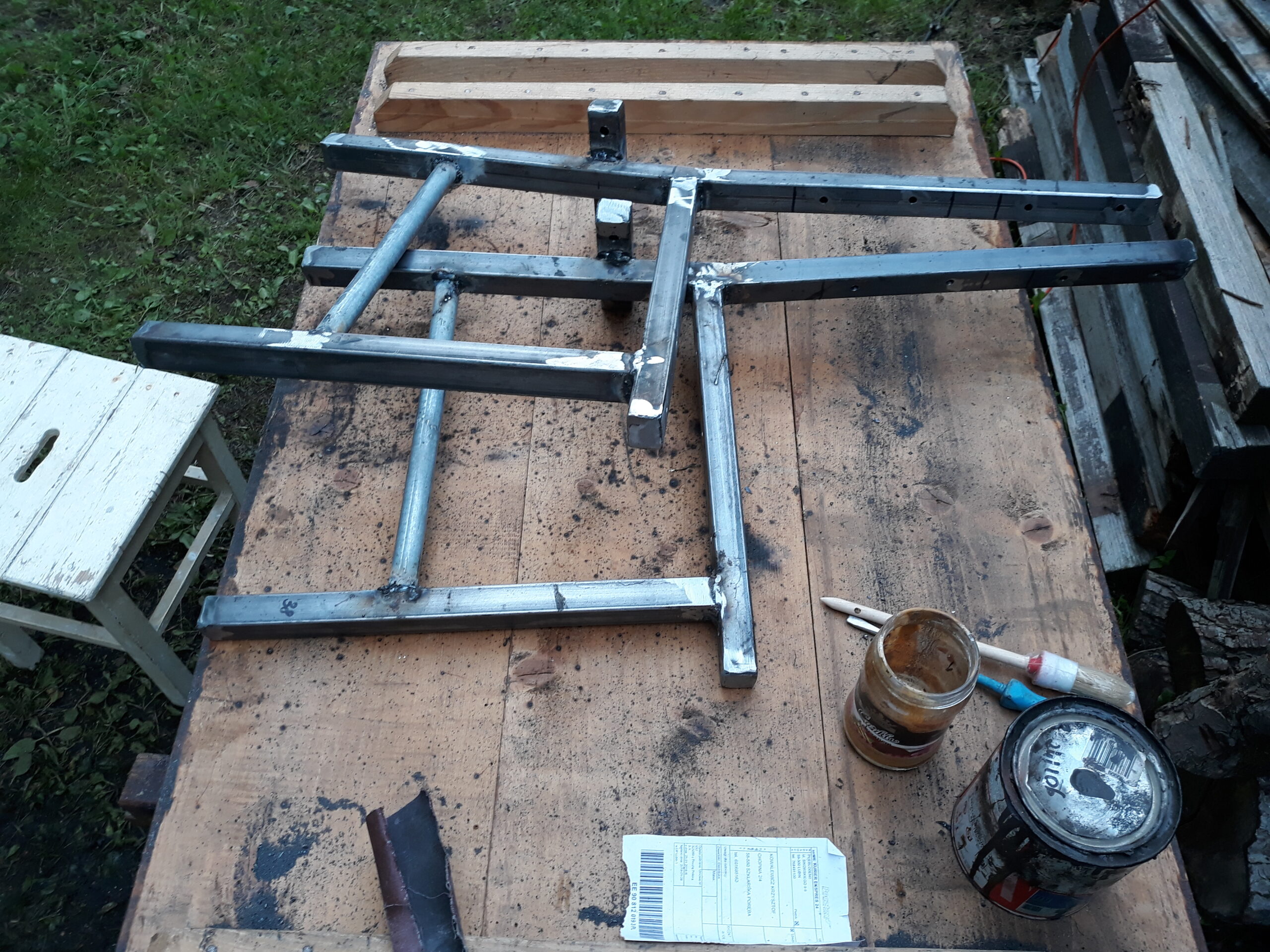 Spawana metalowa konstrukcja ławki ułożona na stole roboczym, z widocznymi elementami ramy, narzędziami oraz materiałami używanymi podczas prac montażowych.