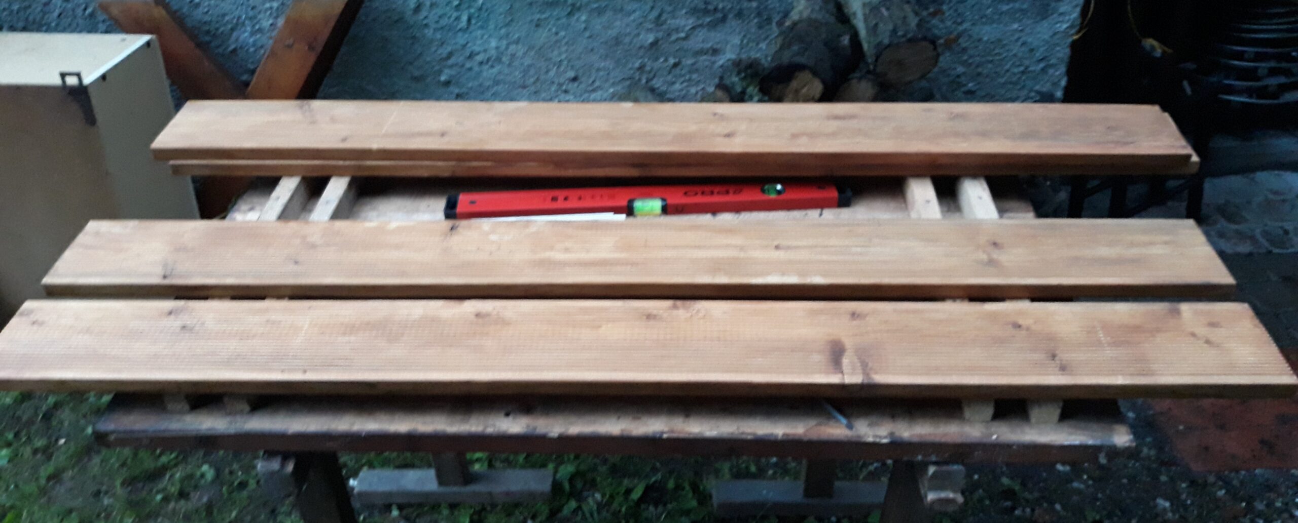 Drewniane deski ułożone na stole roboczym z widoczną poziomicą, przygotowane do montażu siedziska i oparcia ławki.