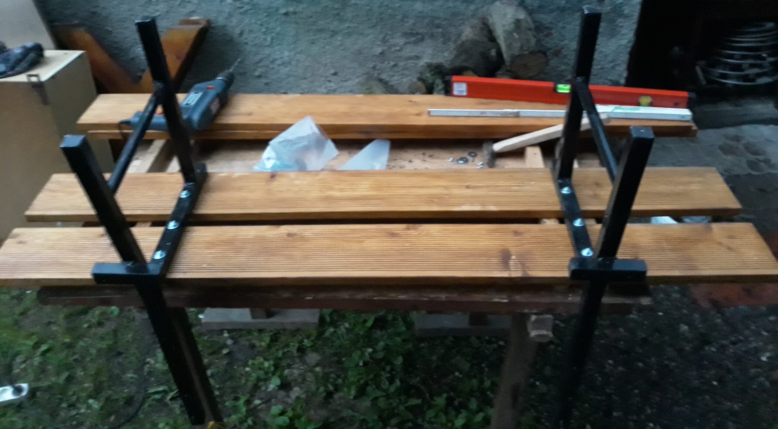 Drewniane deski siedziska przykręcone do metalowych nóg ławki na stanowisku roboczym, pokazujące etap składania konstrukcji.