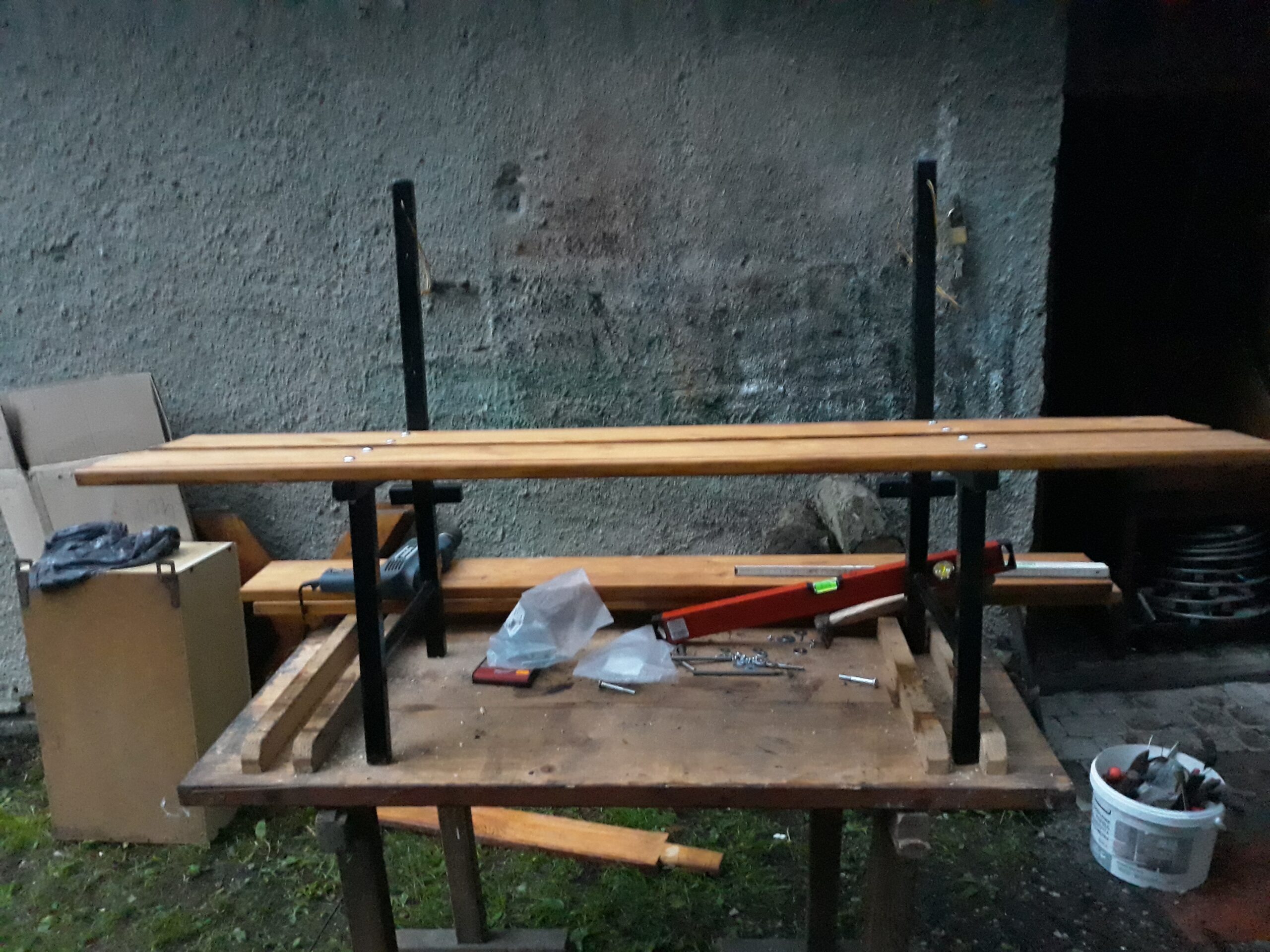 Drewniane elementy ławki zamocowane do metalowej konstrukcji na stole roboczym, przedstawiające etap montażu siedziska i oparcia.