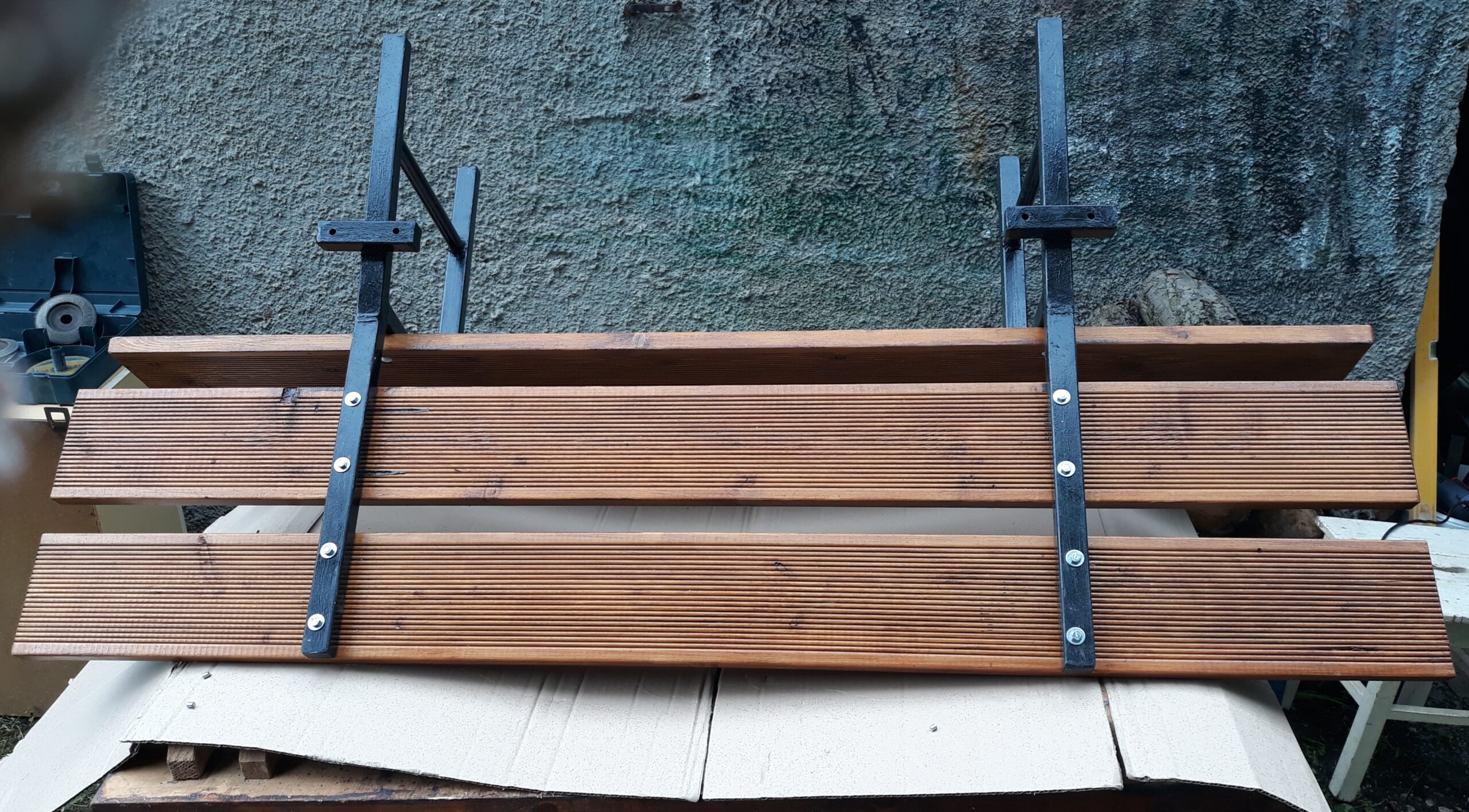 Drewniane siedzisko ławki z zamontowanymi metalowymi wspornikami, ułożone na kartonie i przygotowane do dalszego montażu lub instalacji.