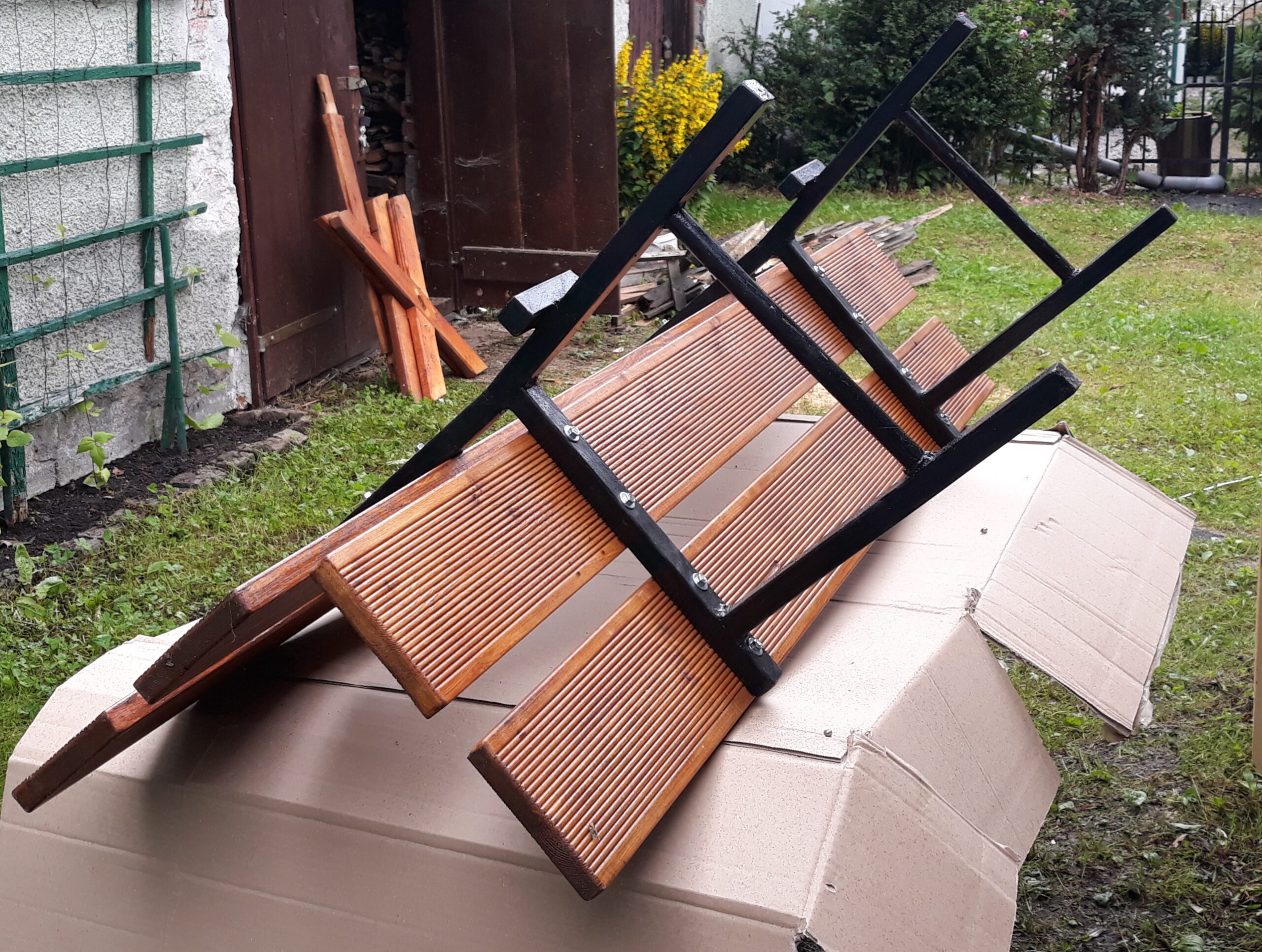Elementy drewnianej ławki z metalową konstrukcją ułożone na kartonie na trawie, przygotowane do montażu lub ponownego ustawienia.