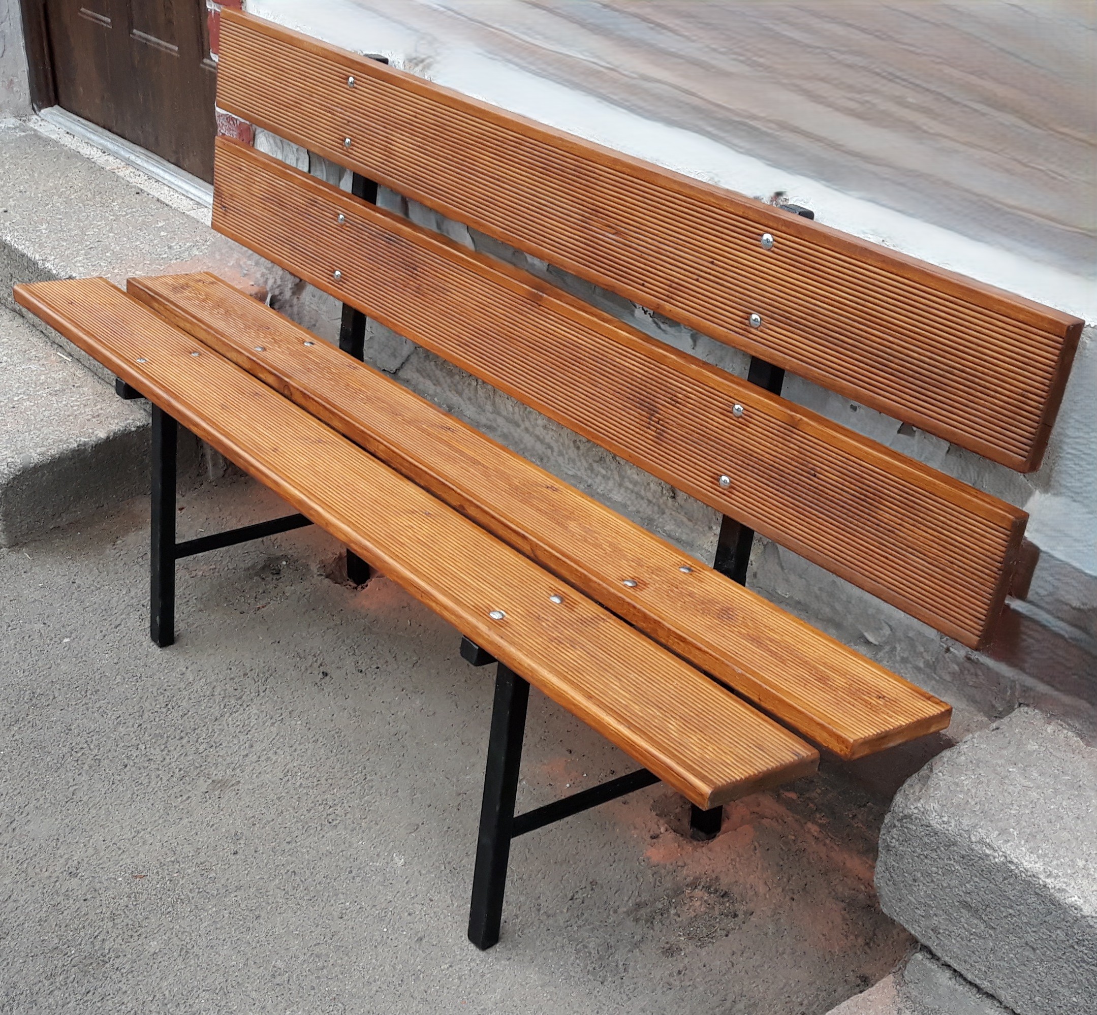 Solidna ławka z drewnianych desek na metalowej konstrukcji, ustawiona przy budynku jako funkcjonalny element małej architektury.